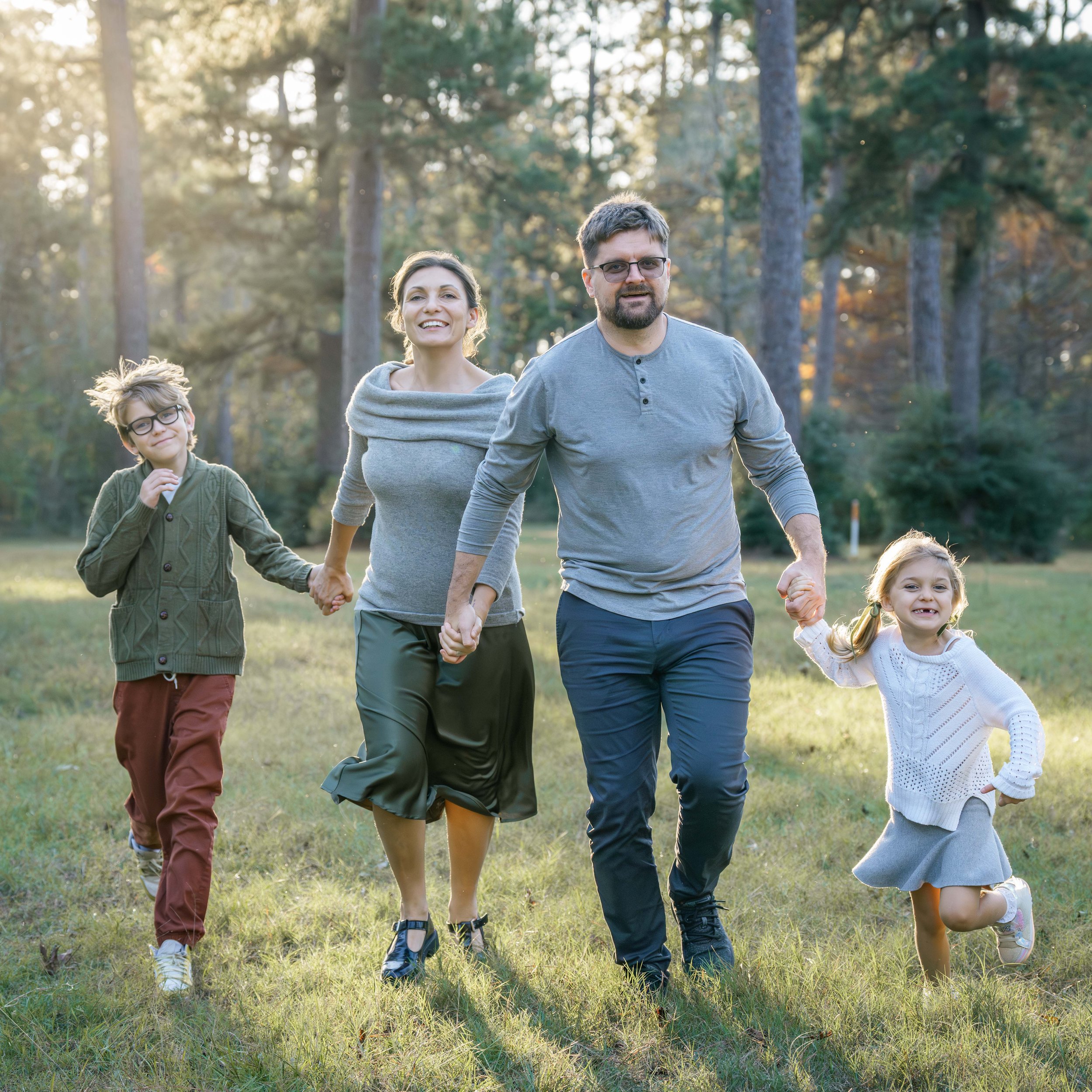 Family photography of a family of four walking hand in hand through a park with tall trees during golden hour, capturing natural connection and movement.