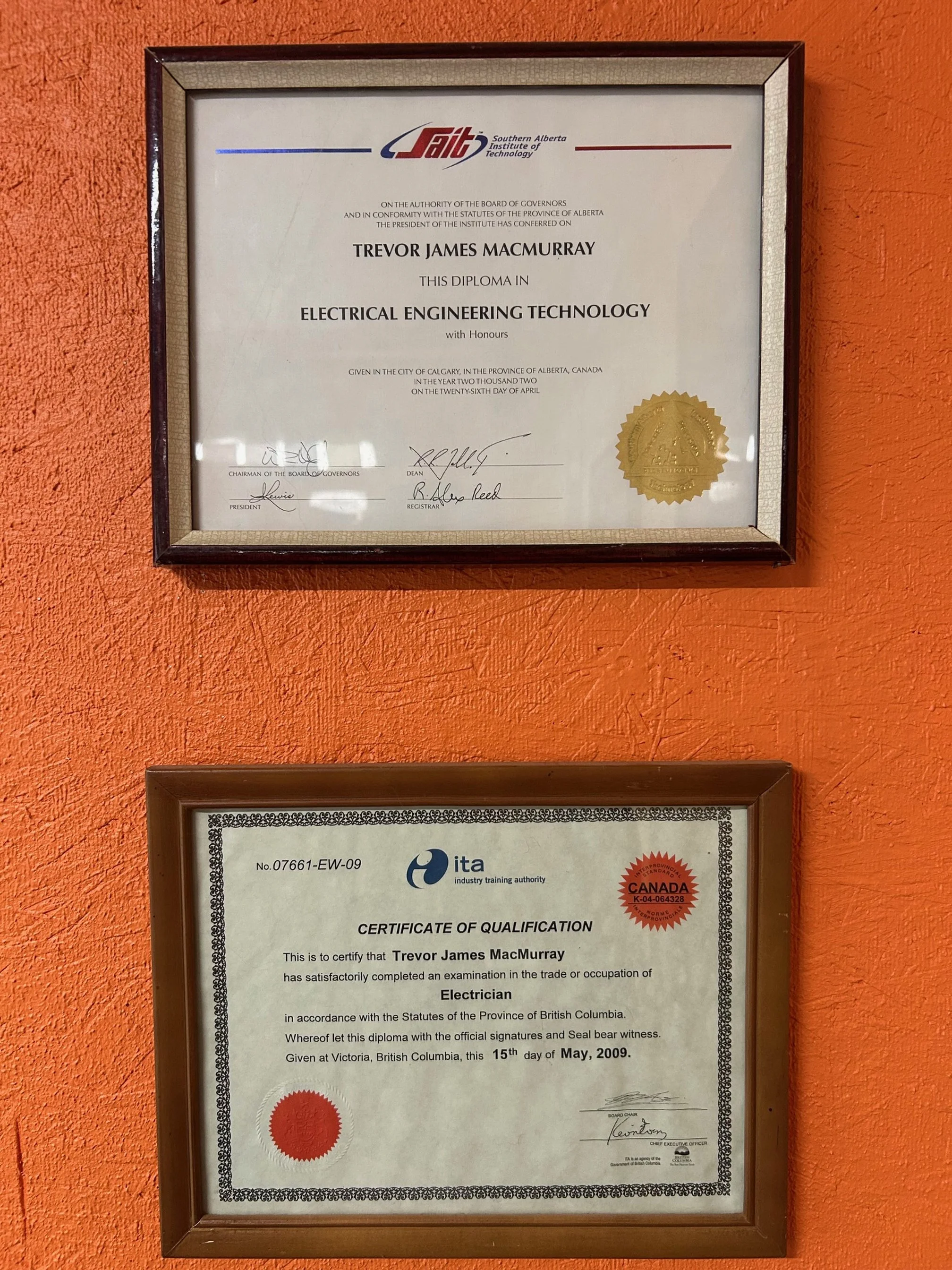 Two framed certificates hanging on an orange wall. The top certificate is from Southern Alberta Institute of Technology, awarded to Trevor James MacMurray for a diploma in Electrical Engineering Technology. The bottom certificate is from industry training authority, certifying Trevor James MacMurray as an electrician, issued in British Columbia on May 15, 2009.