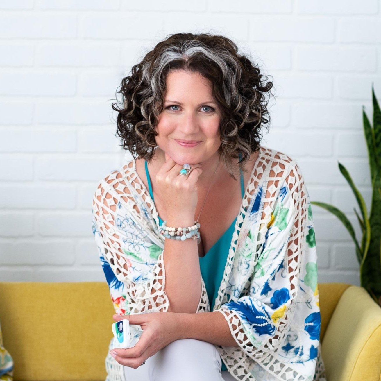 A woman with curly hair and blue eyes smiling and resting her chin on her hand, sitting on a yellow couch with a potted plant nearby.