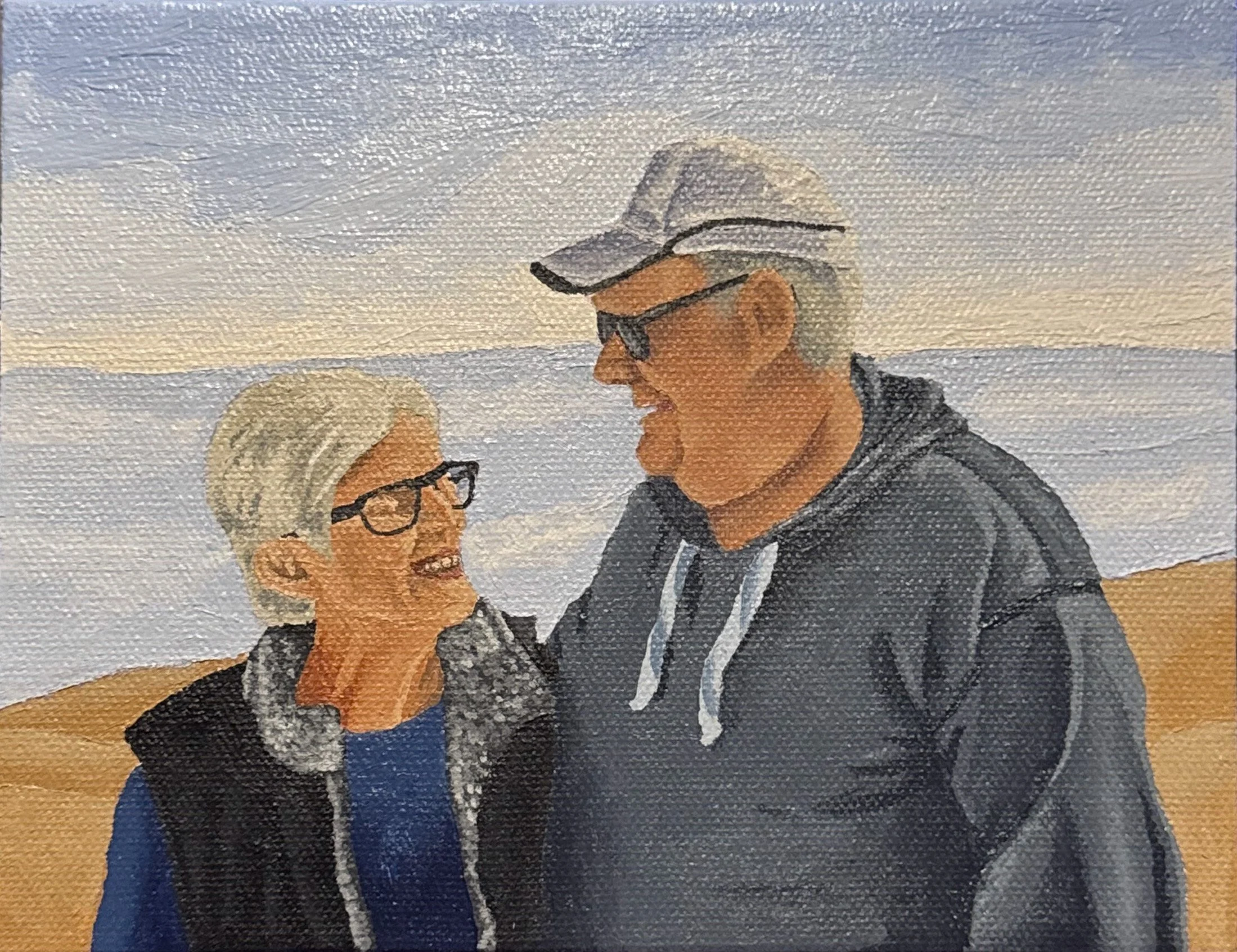 An elderly couple with glasses smiling at each other on a beach with the ocean and sky in the background.