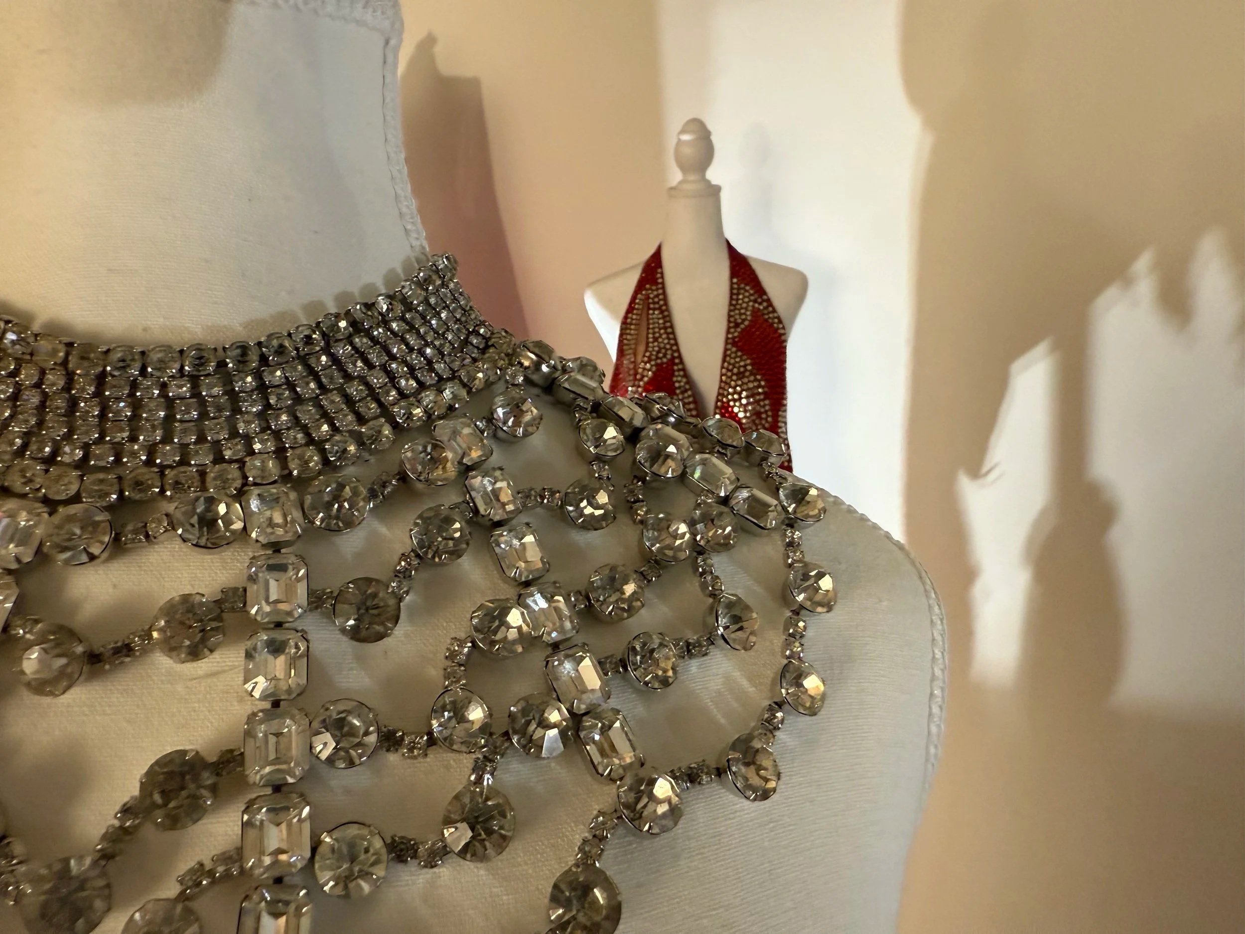 Display of jewelry including rhinestone necklaces, a red and gold piece, and a small dress form on a white mannequin with scattered jewelry on a platform.