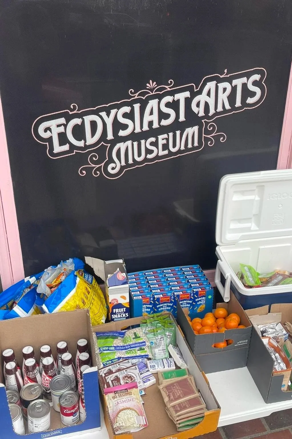 Table with assorted snacks and food items in front of a blackboard with the text 'Eccy Sia Arts Museum'.