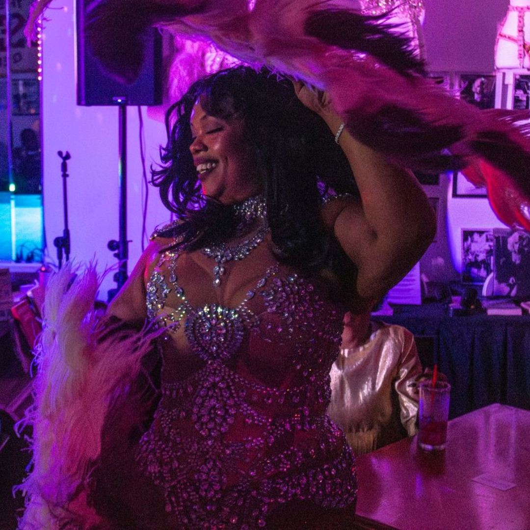 A woman dancing at a party, wearing a sparkling, jewel-encrusted outfit with feather accessories, lit with purple and pink lighting.