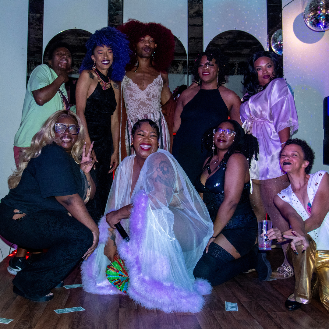 Group of diverse women celebrating at a party, some making peace signs, others smiling broadly, dressed in colorful and stylish outfits, with disco balls and reflective décor in the background.