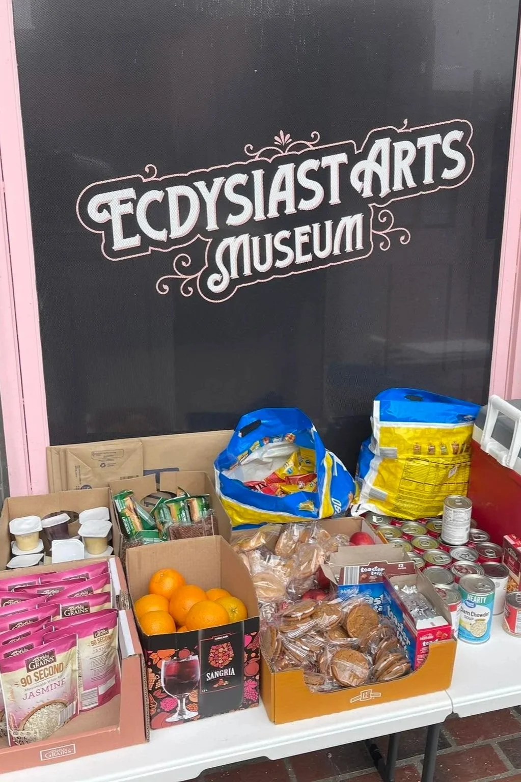 Table with snacks and canned goods in front of a blackboard with the sign 'Ecstasy Arts Museum'.