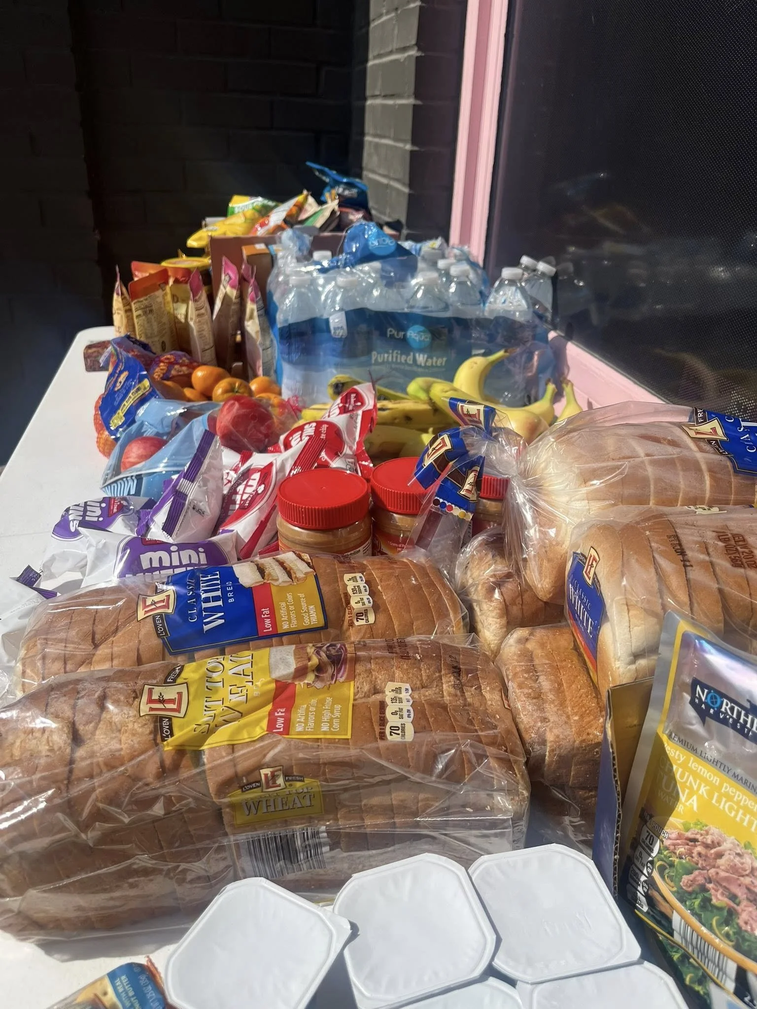 Grocery items on a white table including bread, bottled water, bananas, oranges, snack bags, peanut butter, and a boxed meal.