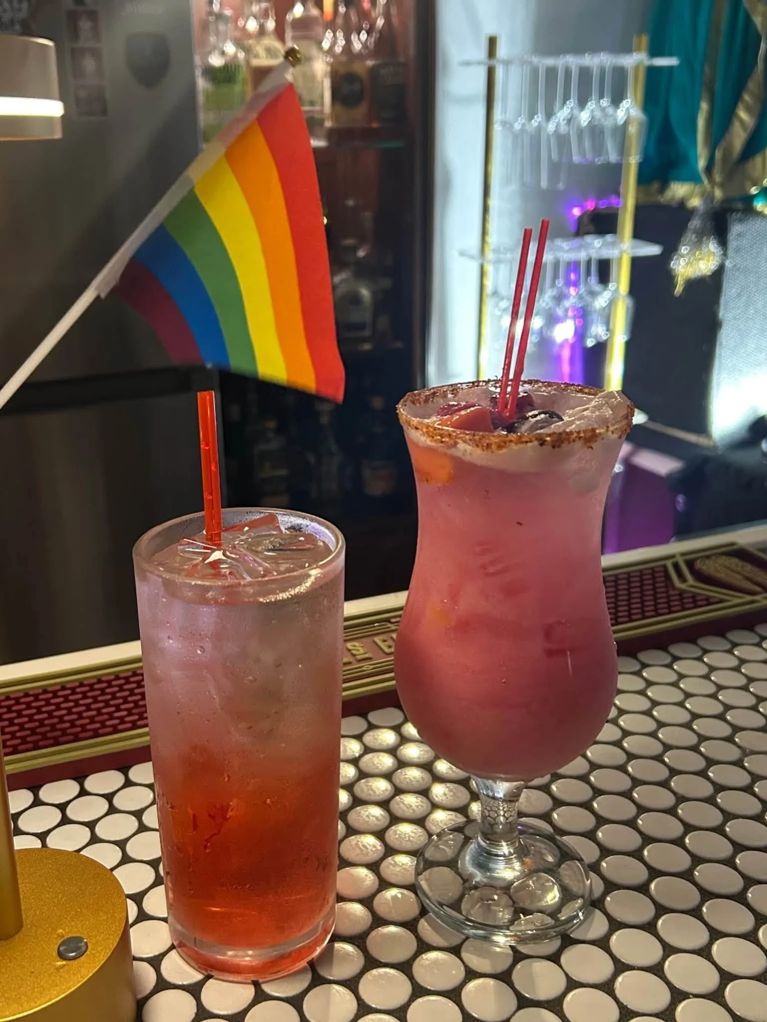 Two pink cocktails with ice and garnishes, one in a tall glass and the other in a hurricane glass, with a small rainbow pride flag behind them, on a bar counter with a black and white polka dot pattern.