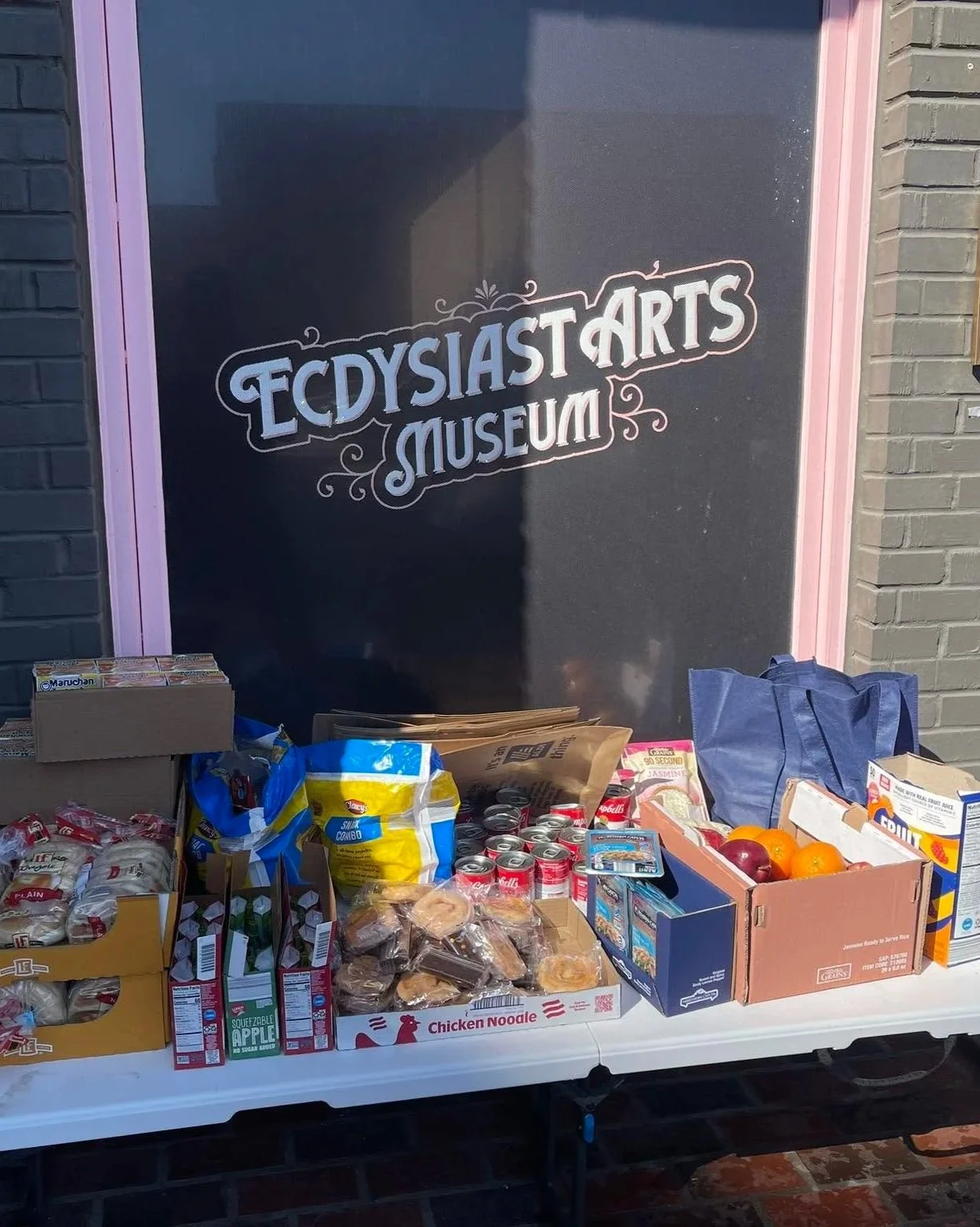Table of groceries including bread, chips, canned drinks, apples, and boxes of food, in front of a sign reading 'Ecdysia Arts Museum' on a brick wall.