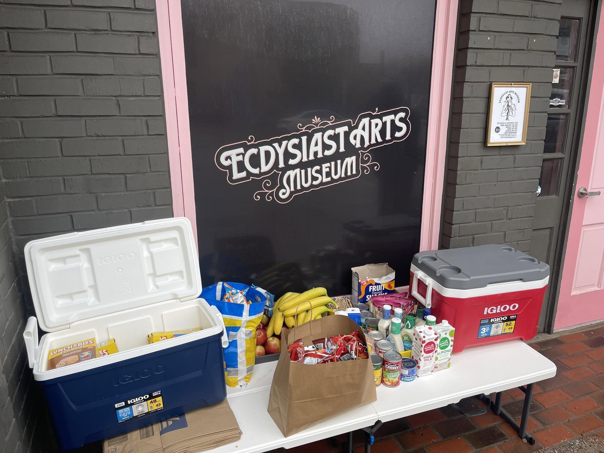 Outside of a building with a sign for the Ecstasy Arts Museum. Several large coolers, food boxes, and canned foods are arranged on a table and on the ground, including bananas, apples, milk, and various canned goods.