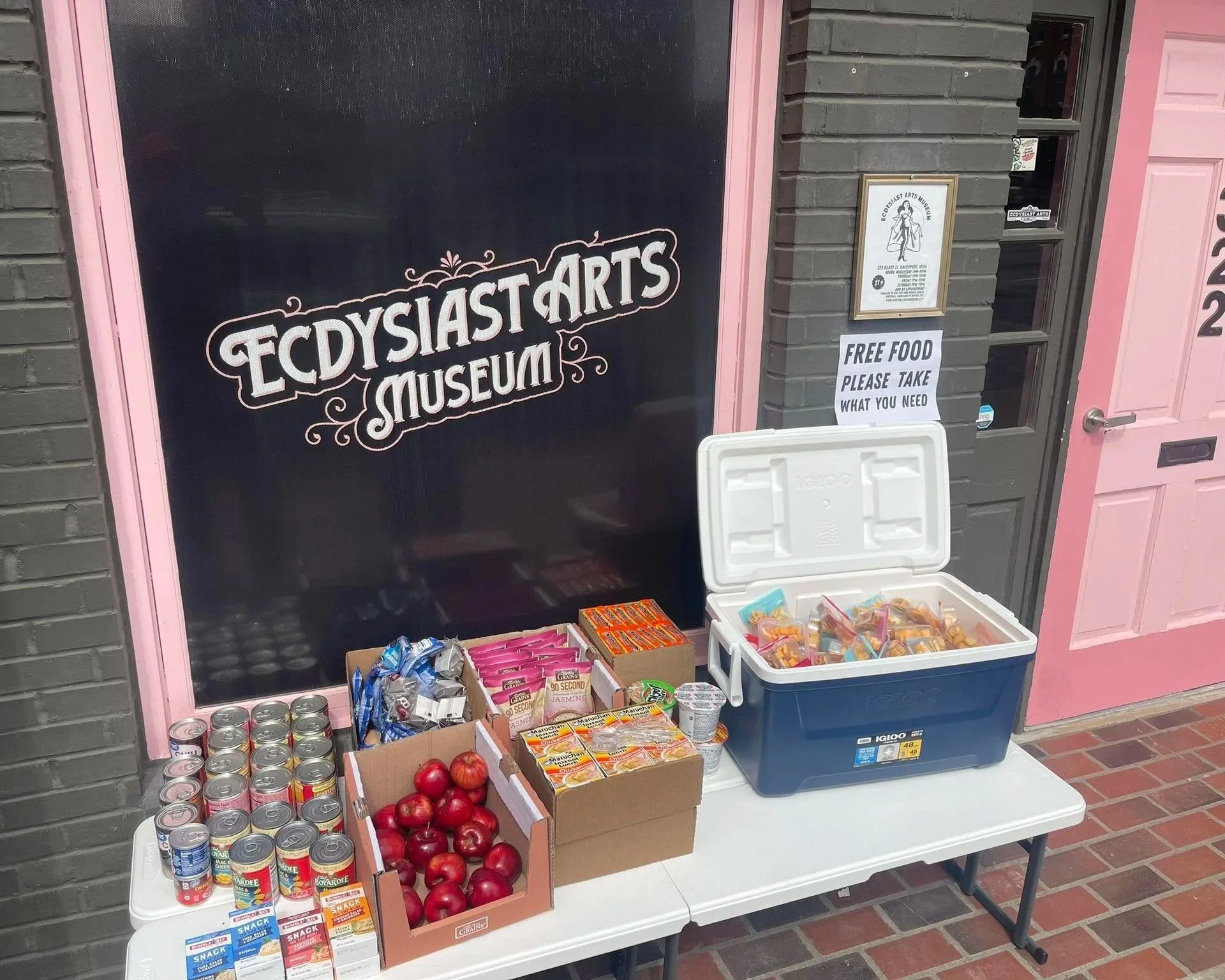 Table set up outside a building with signs offering free food, including canned goods, packaged snacks, apples, and candies, in front of a black and pink building with a framed poster.