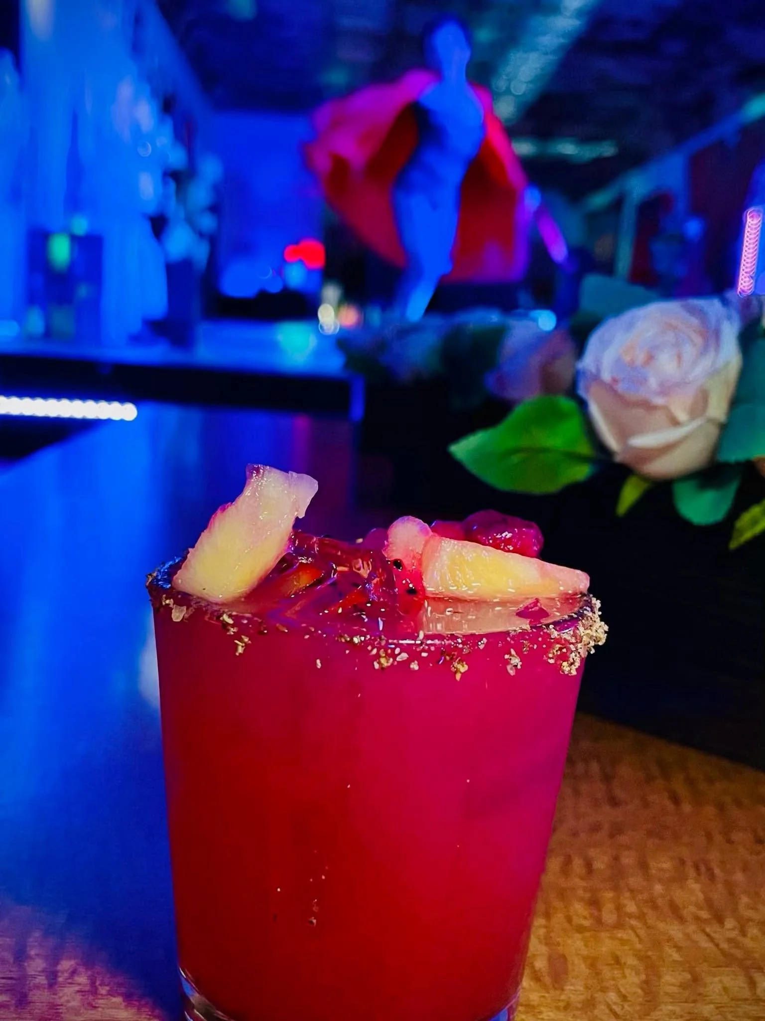Pink cocktail glass filled with a drink garnished with pineapple slices and a cherry, with a salted rim, in a dimly lit environment with blue and purple lighting.