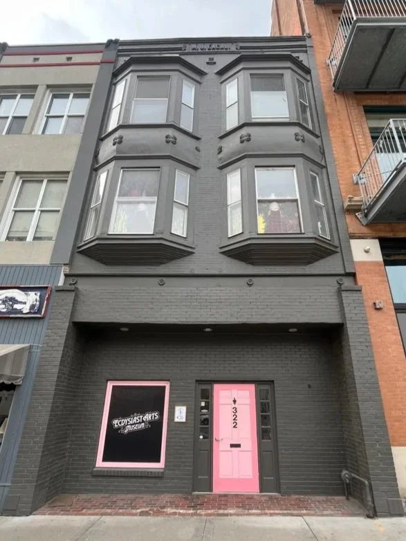 The building has a black facade with a pink door and a large window with a pink border, displaying the words 'ECDYSIAST ARTS MUSEUM' in white. The upper floors have protruding bay windows showcasing our upcoming vintage shop space.