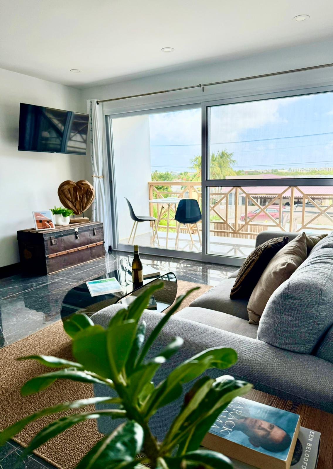 Living room with a cream sofa, a black glass coffee table with magazines, a wall-mounted flat screen TV, and sliding glass doors leading to a balcony with outdoor furniture. Outside, there are trees and a partly cloudy sky.