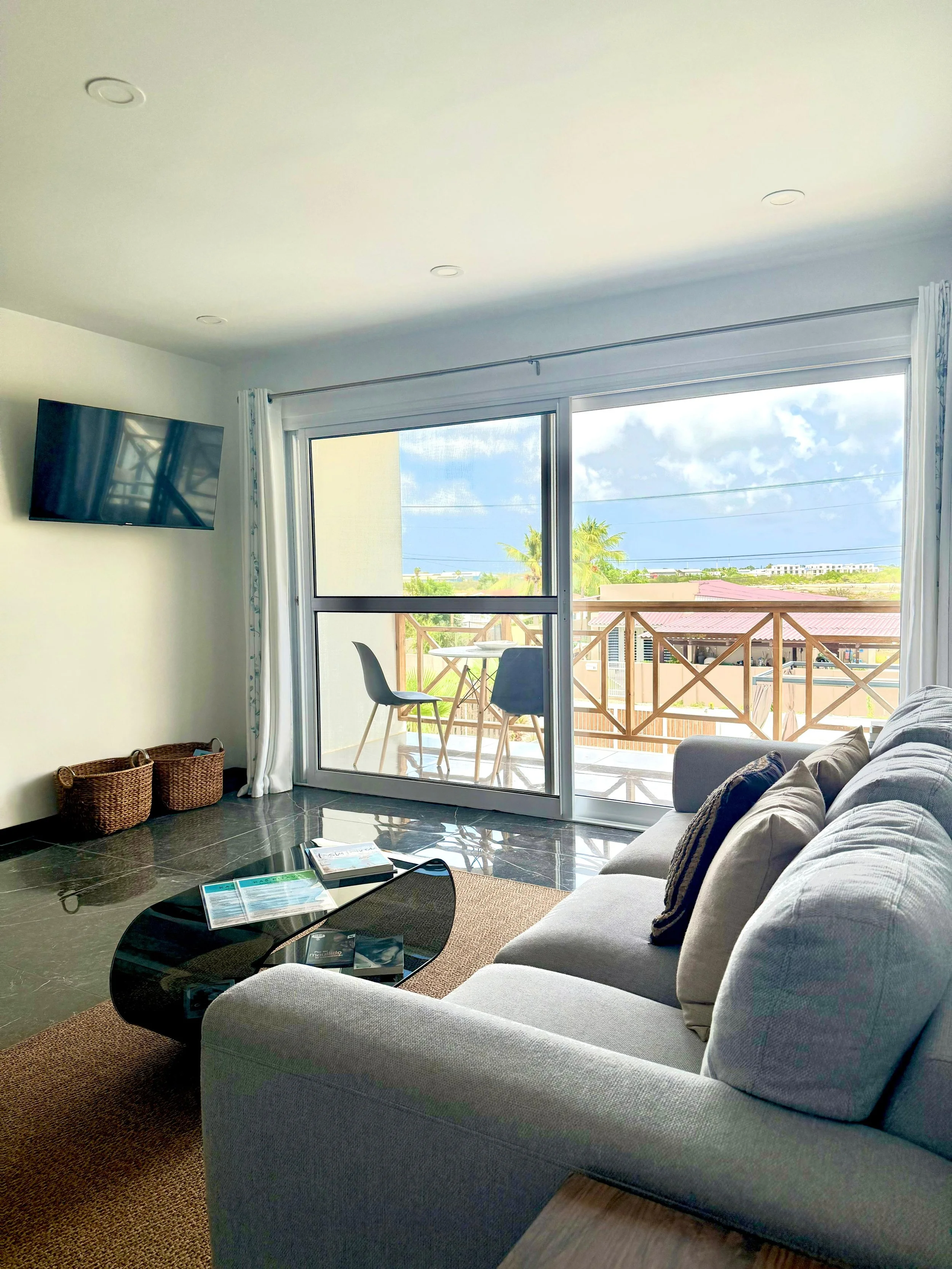 Living room with a cream sofa, a black glass coffee table with magazines, a wall-mounted flat screen TV, and sliding glass doors leading to a balcony with outdoor furniture. Outside, there are trees and a partly cloudy sky.