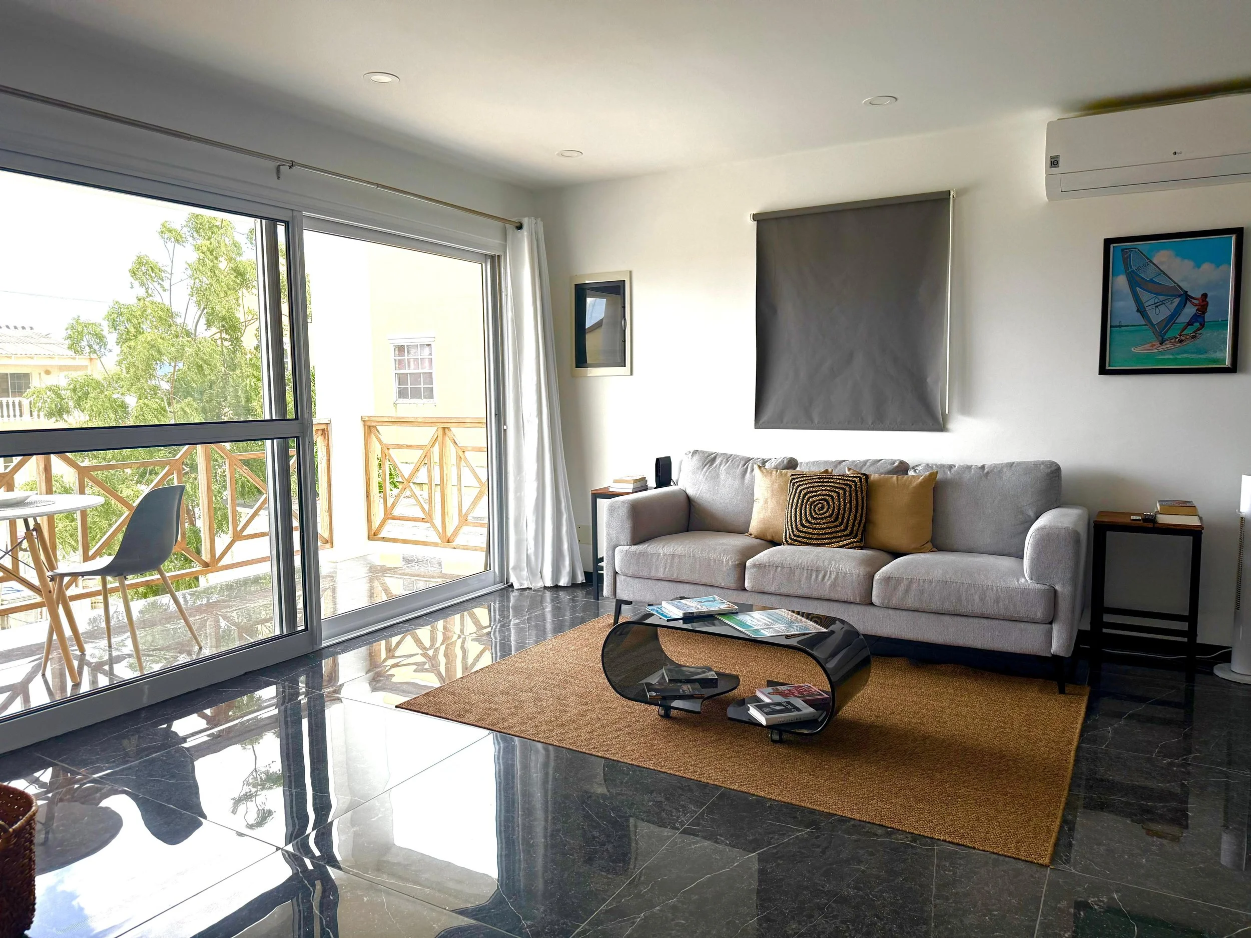 Living room with a large sliding glass door leading to a balcony, a gray sofa with two pillows, a black and brown coffee table, a tan rug, two side tables with books, framed artwork on the walls, and an air conditioning unit on the wall.