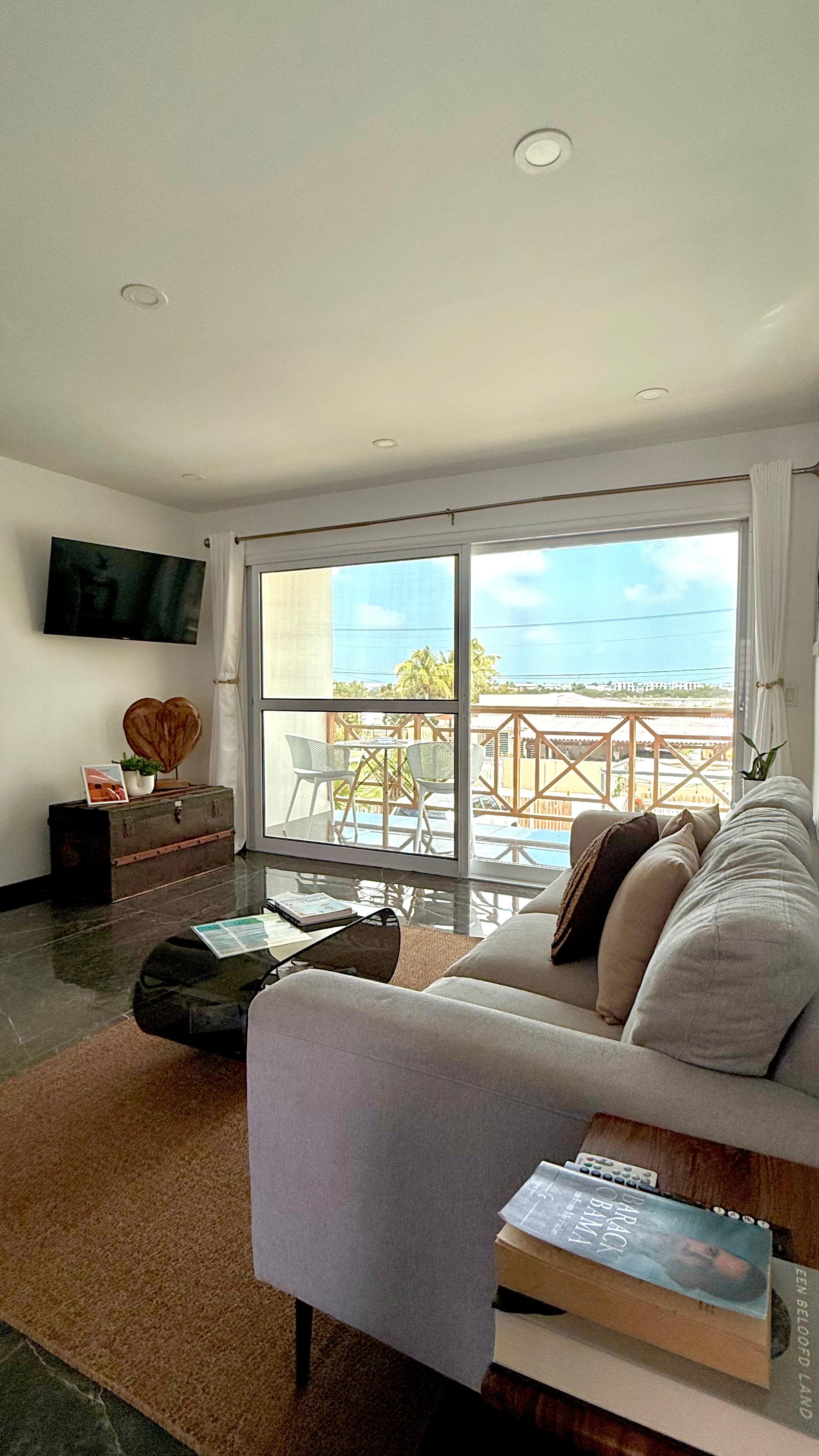Living room with a cream sofa, a black glass coffee table with magazines, a wall-mounted flat screen TV, and sliding glass doors leading to a balcony with outdoor furniture. Outside, there are trees and a partly cloudy sky.