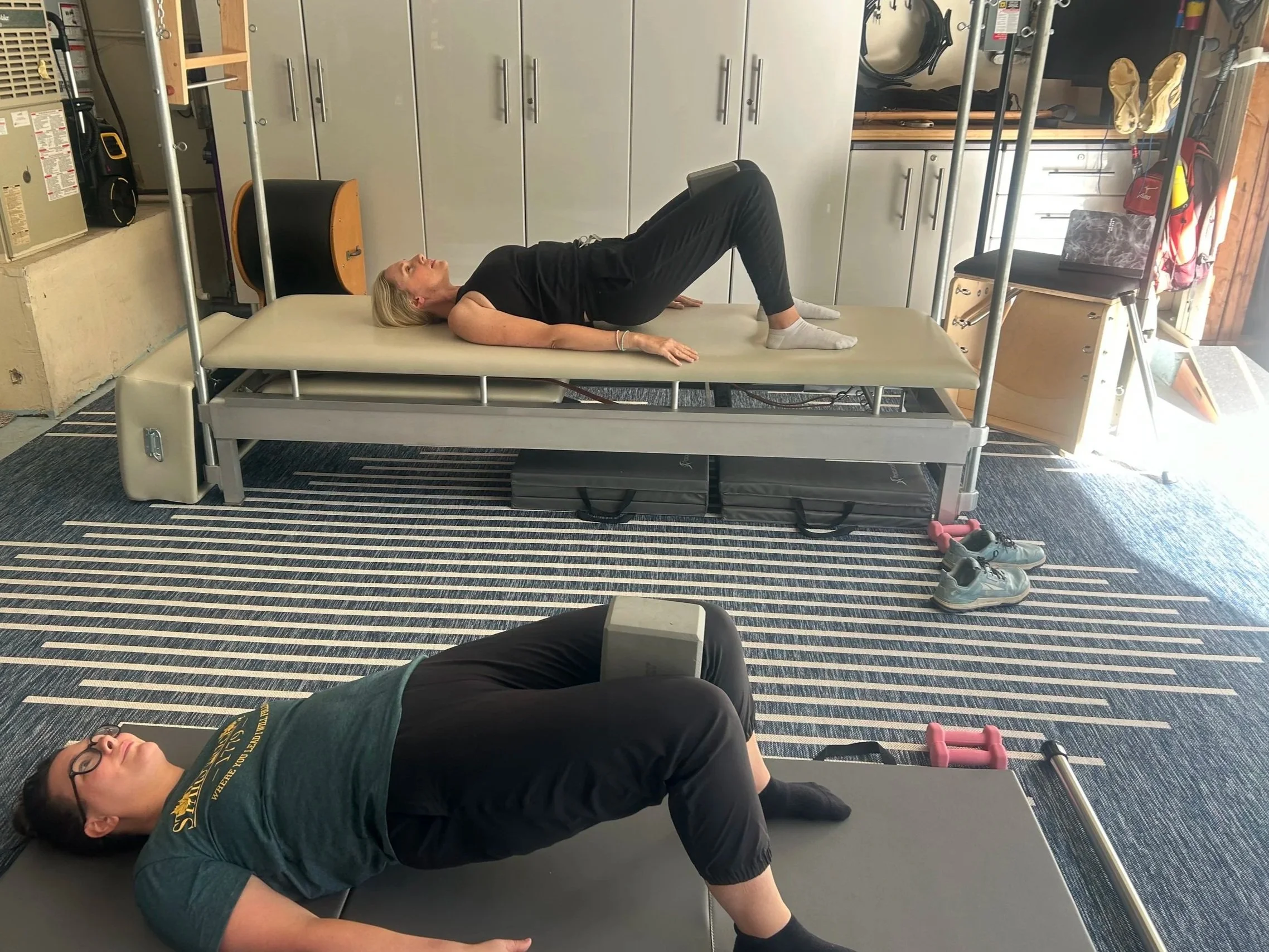 Two women lying on therapy tables, performing physical therapy exercises with weights in a gym or therapy room.