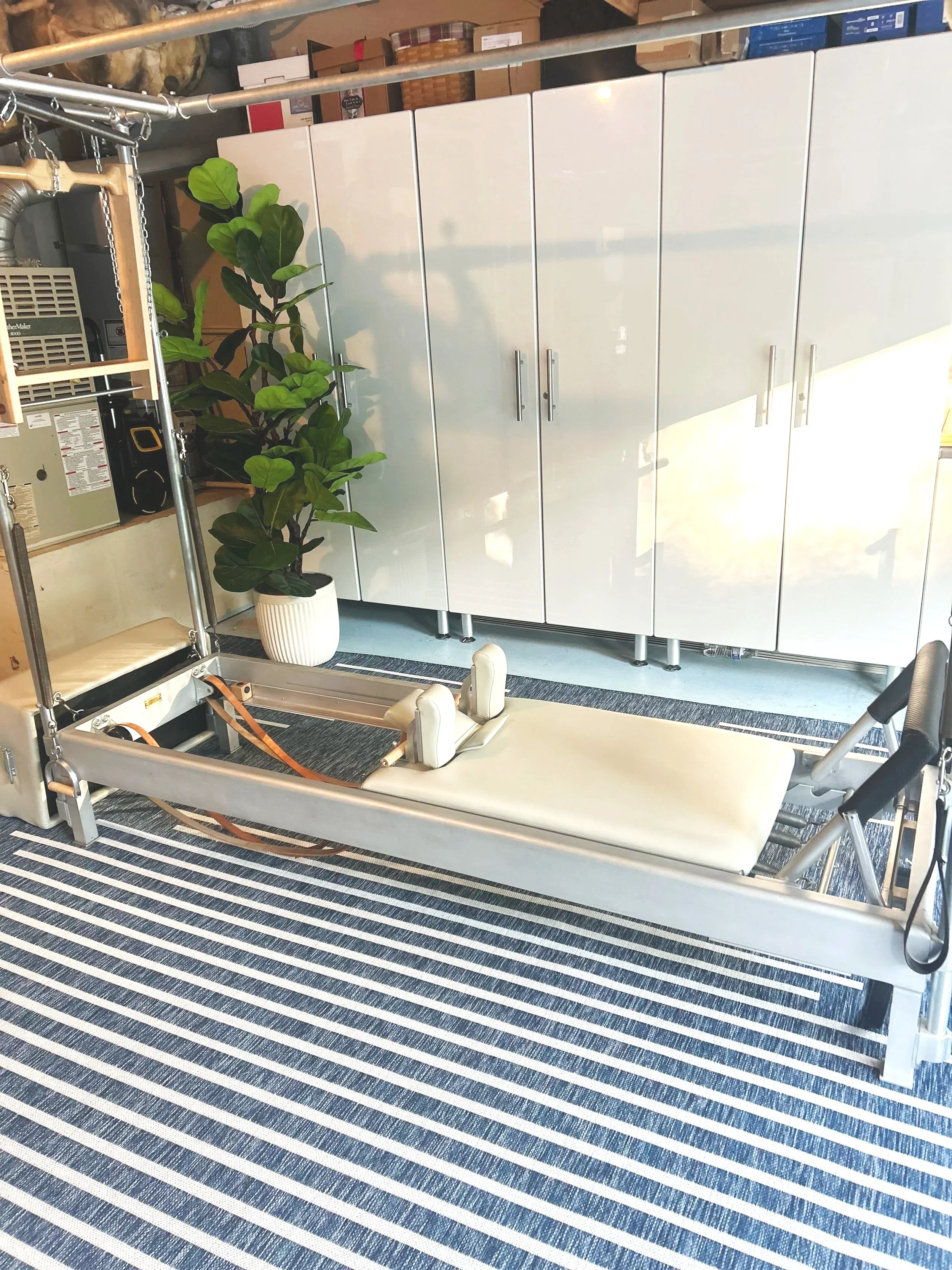A Reformers Recovery Pilates reformer machine in a room with a striped rug, a large potted green plant, and closed white cabinets in the background.