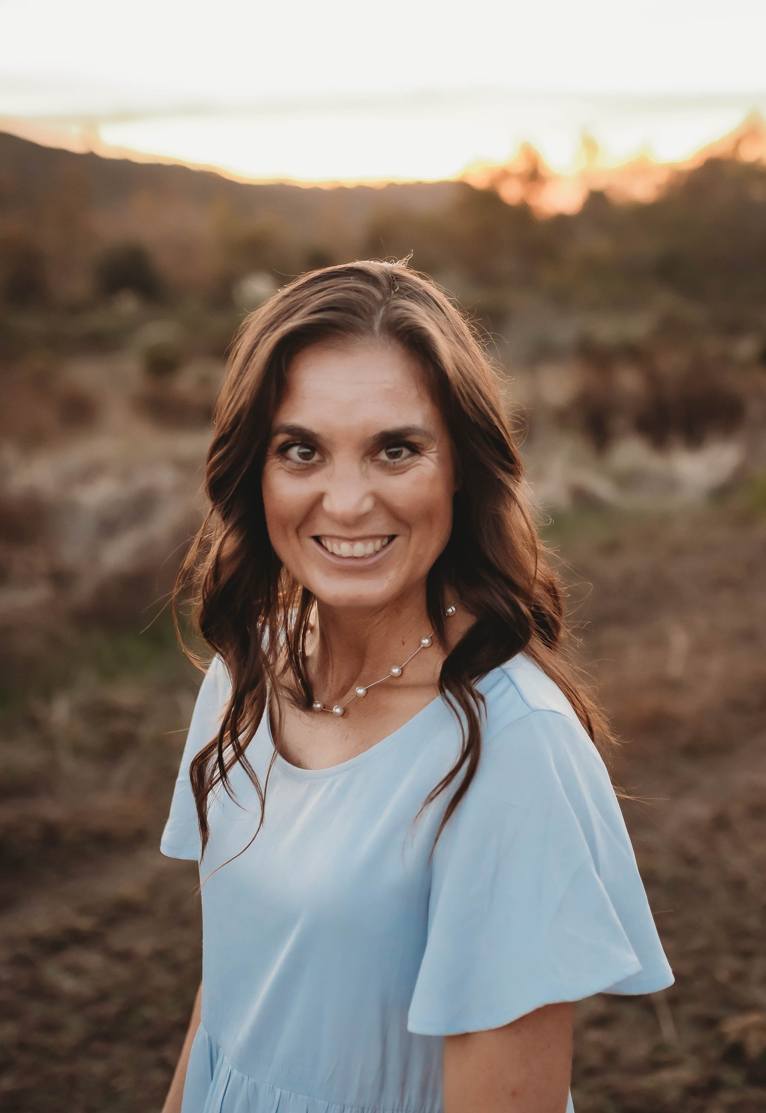A woman smiling outdoors at sunset, wearing a light blue dress and a pearl necklace, with a blurred natural landscape in the background.