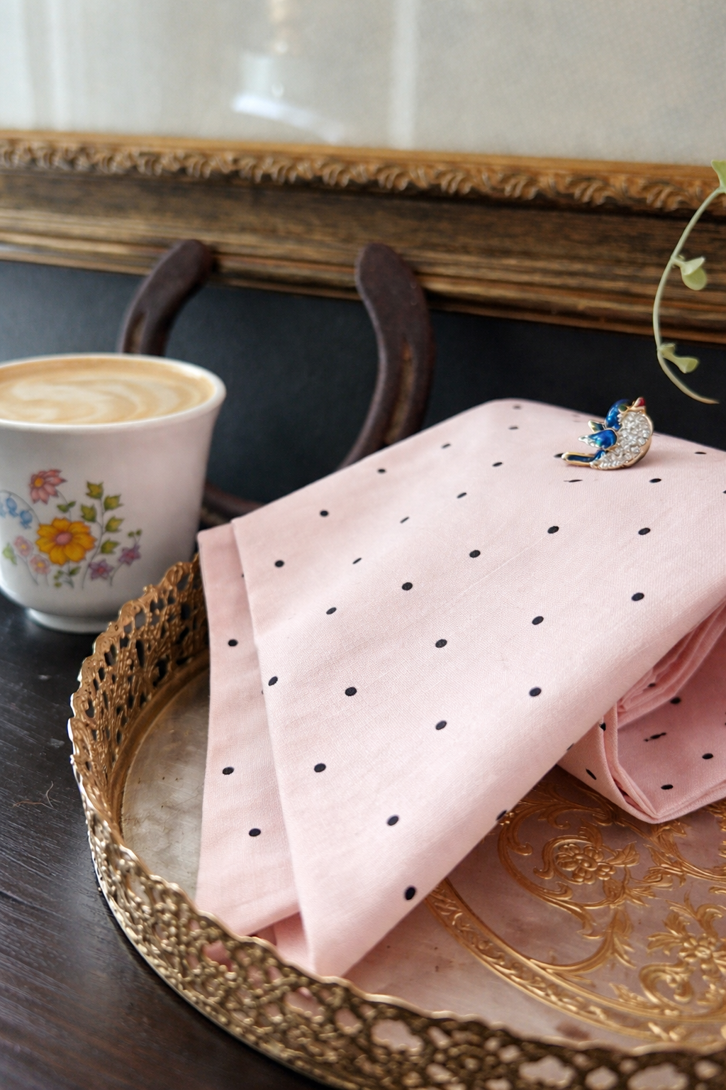 A pink cloth with black polka dots on a decorative gold tray, a pink coffee cup with a floral pattern filled with coffee, and a gemstone brooch shaped like a moon with blue accents.