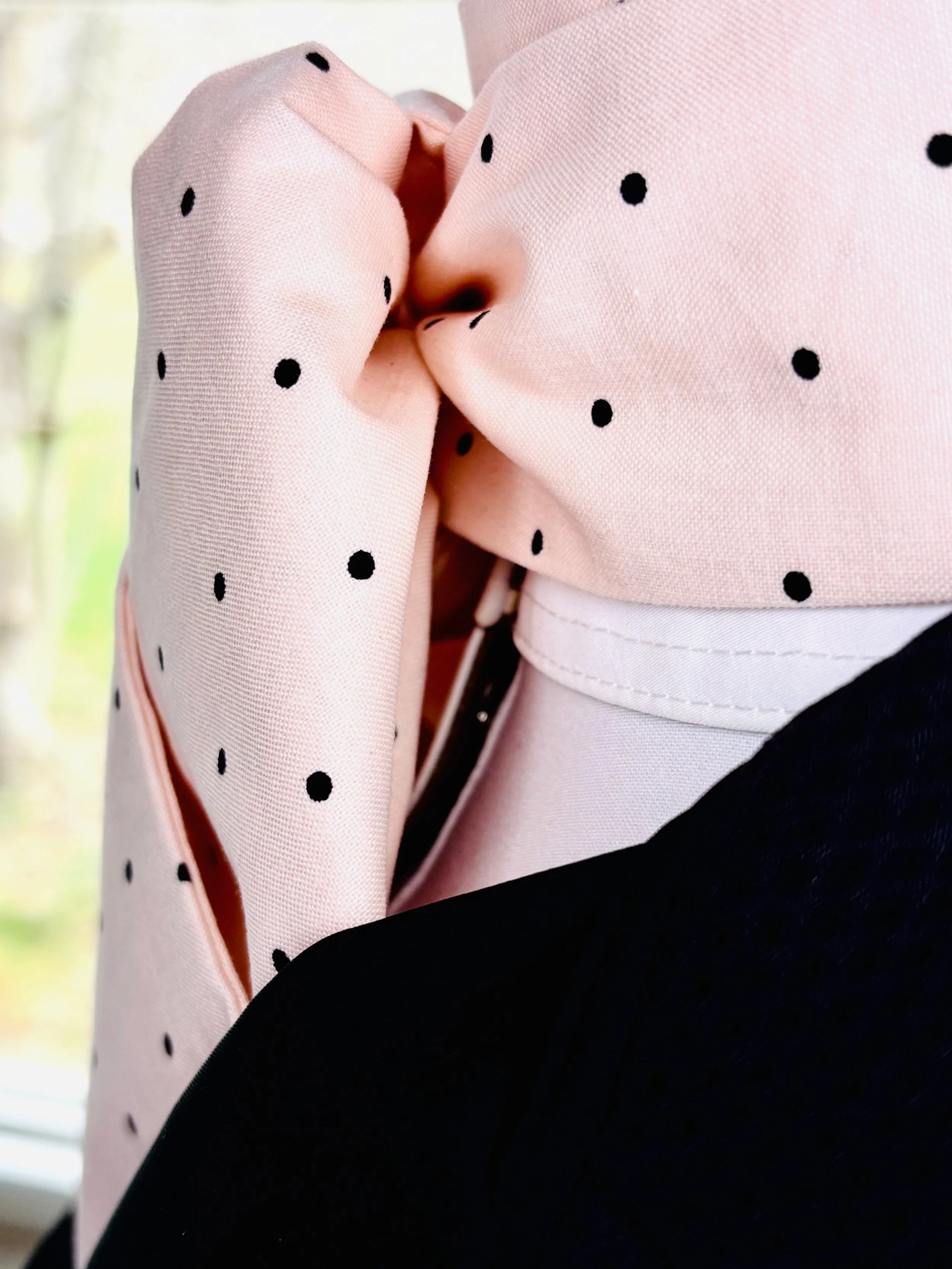 Close-up of a person wearing a pink, polka-dotted fabric with black dots, holding it tightly, with a blurred view outside a window in the background.