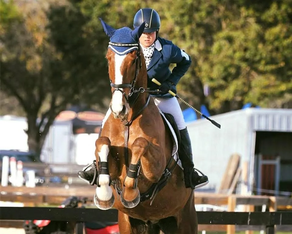 A female equestrian rider wearing a helmet, navy jacket, and white riding pants jumps over a hurdle on a brown and white horse during an outdoor event.