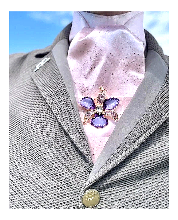 Close-up of a person wearing a pink neck scarf with a butterfly-shaped brooch featuring purple wings and gold accents, over a gray textured blazer.