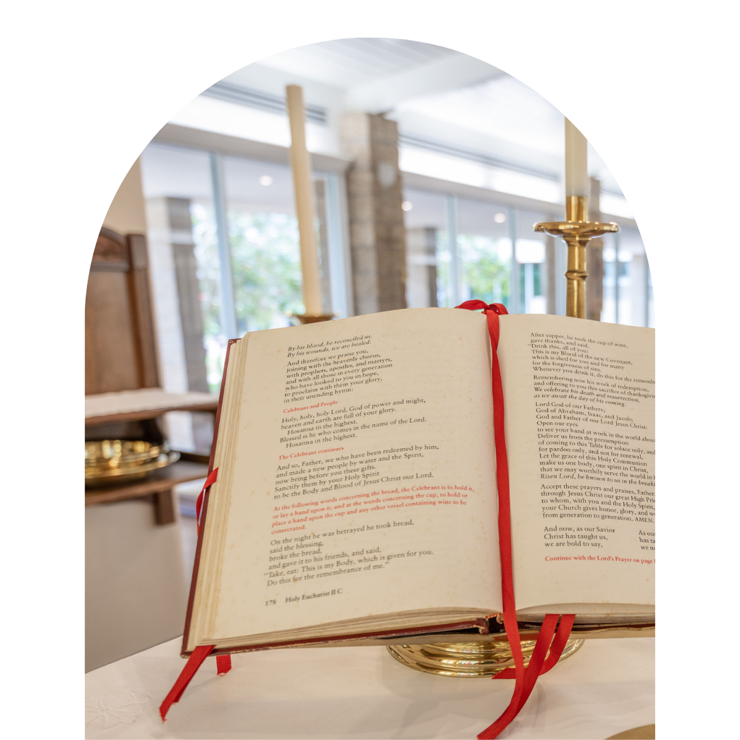 An open Bible on a white table, with candles and a gold-colored stand in the background, inside a church or chapel.