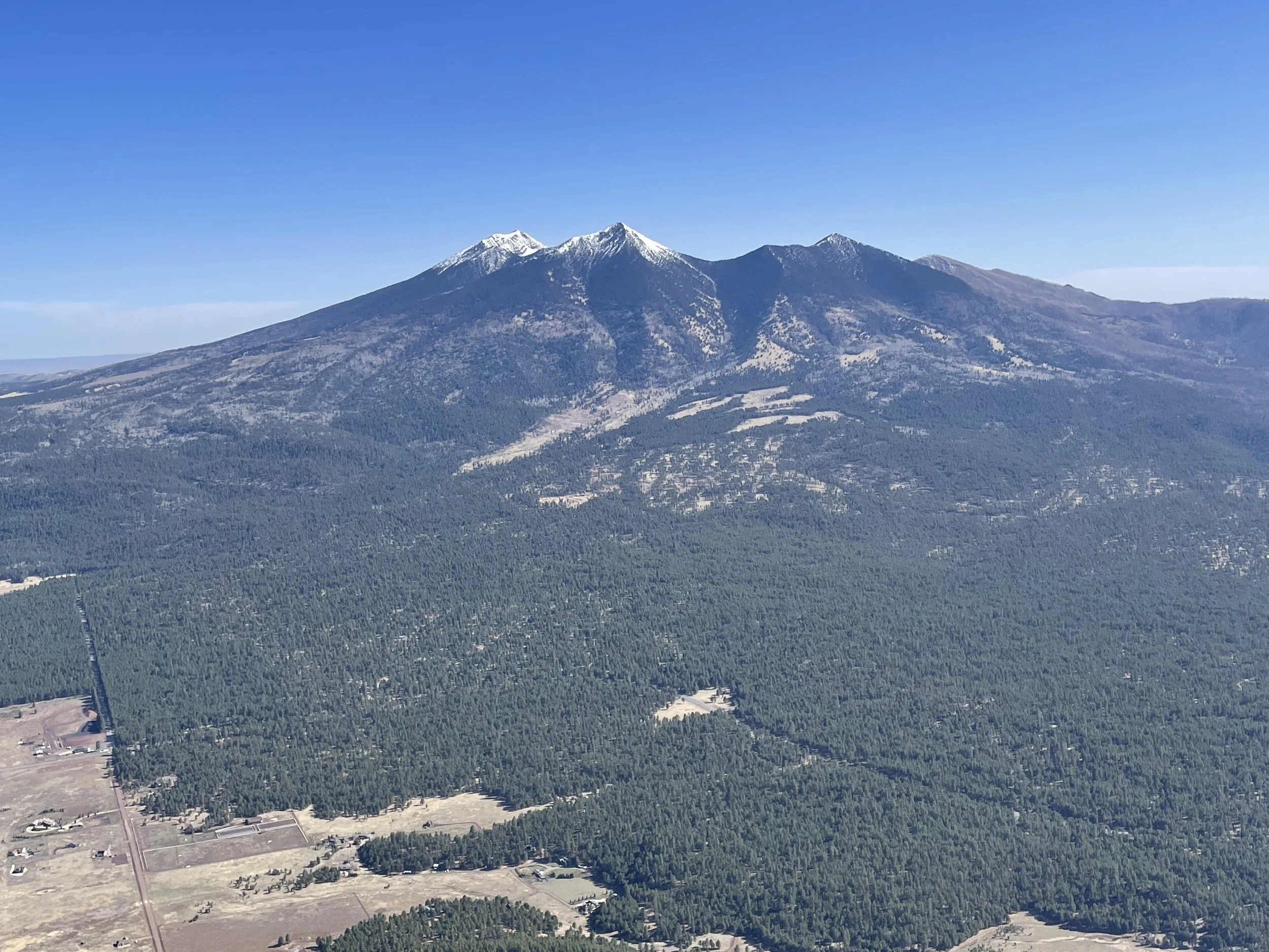 Aerial photograph of a mountain with snow-capped peaks and forested slopes, with a small settlement and fields in the foreground. Bluebird Air Tours