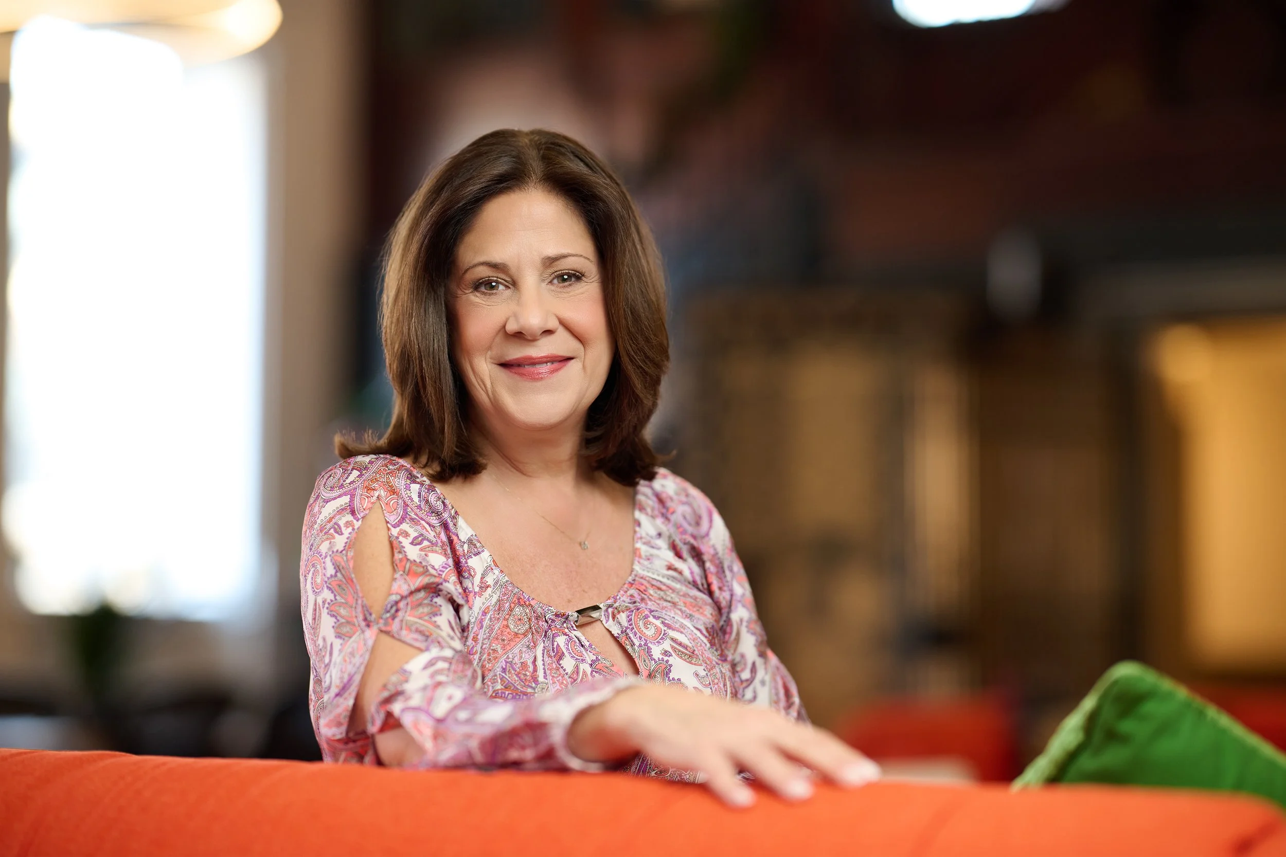 A woman with shoulder-length brown hair smiling while resting her hand on a red couch in a warmly lit living room.