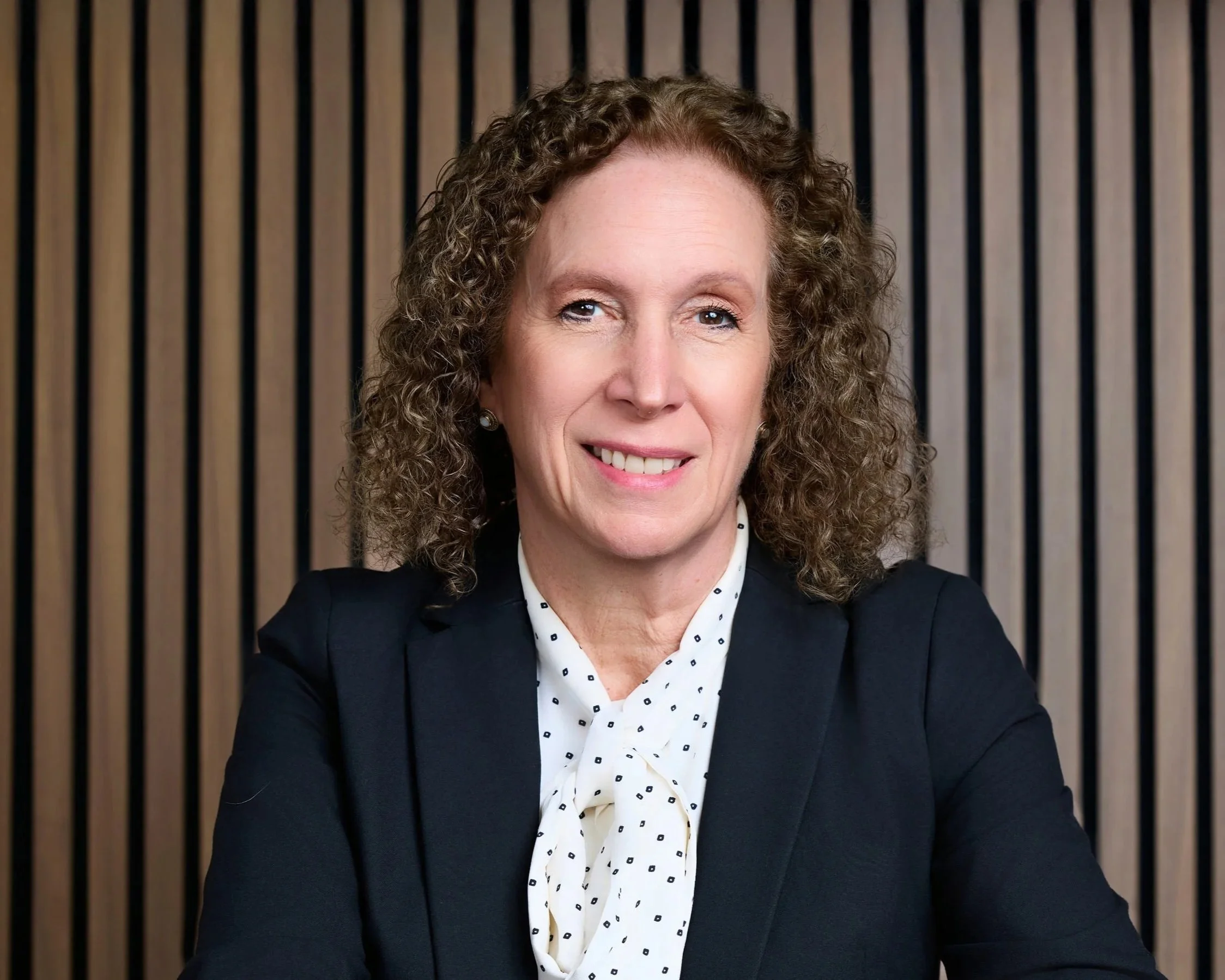 A woman with curly brown hair wearing a black blazer and polka dot blouse, smiling, in front of a vertical wooden slat background.