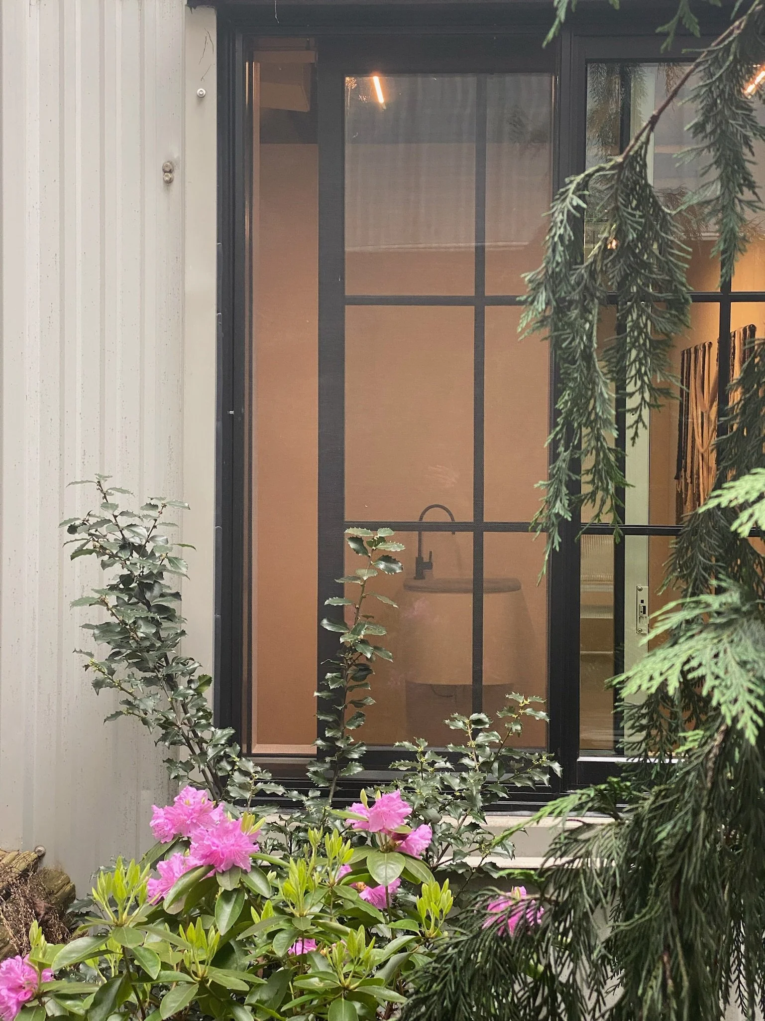 View of a screened porch with a closed door, plants, and pink flowers in the foreground.