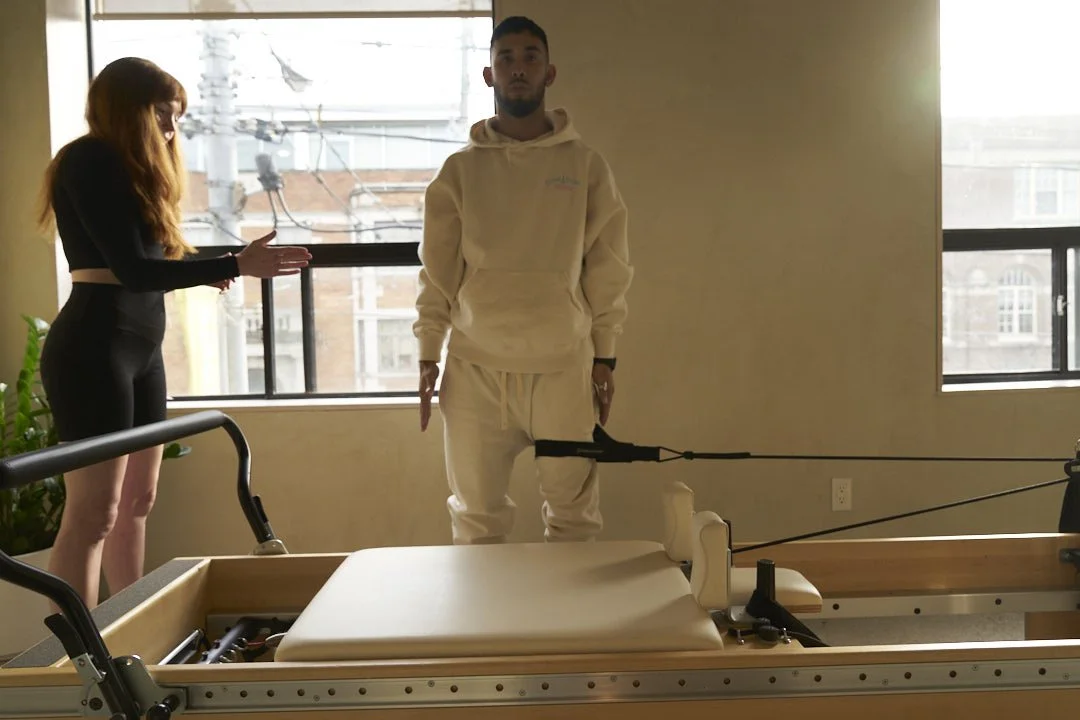 A man in a white hoodie and pants standing next to a therapy or rehabilitation table with an arm attachment, while a woman with long hair in a black outfit gestures towards him, in a room with large windows.