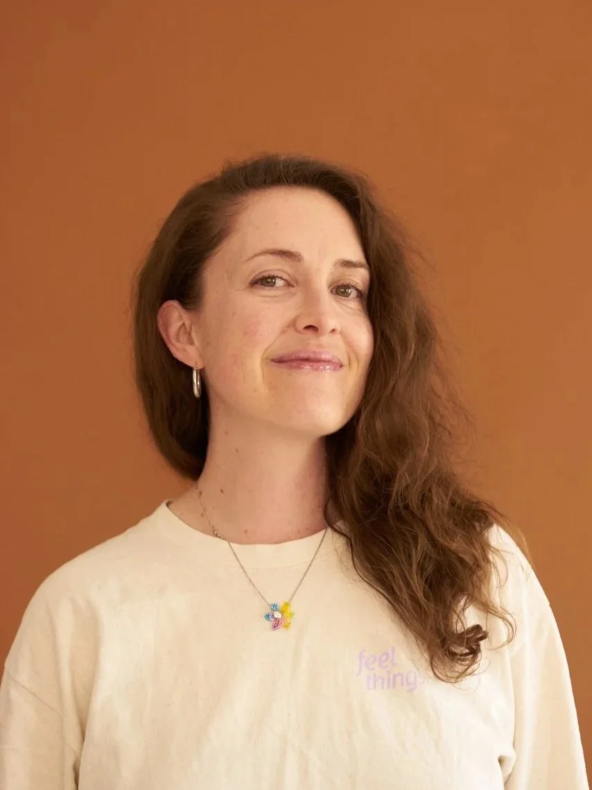 A woman with medium red hair wearing a cream-colored T-shirt with the words 'feel things' partially visible, a colorful butterfly necklace, and hoop earrings, standing against a solid brown background.