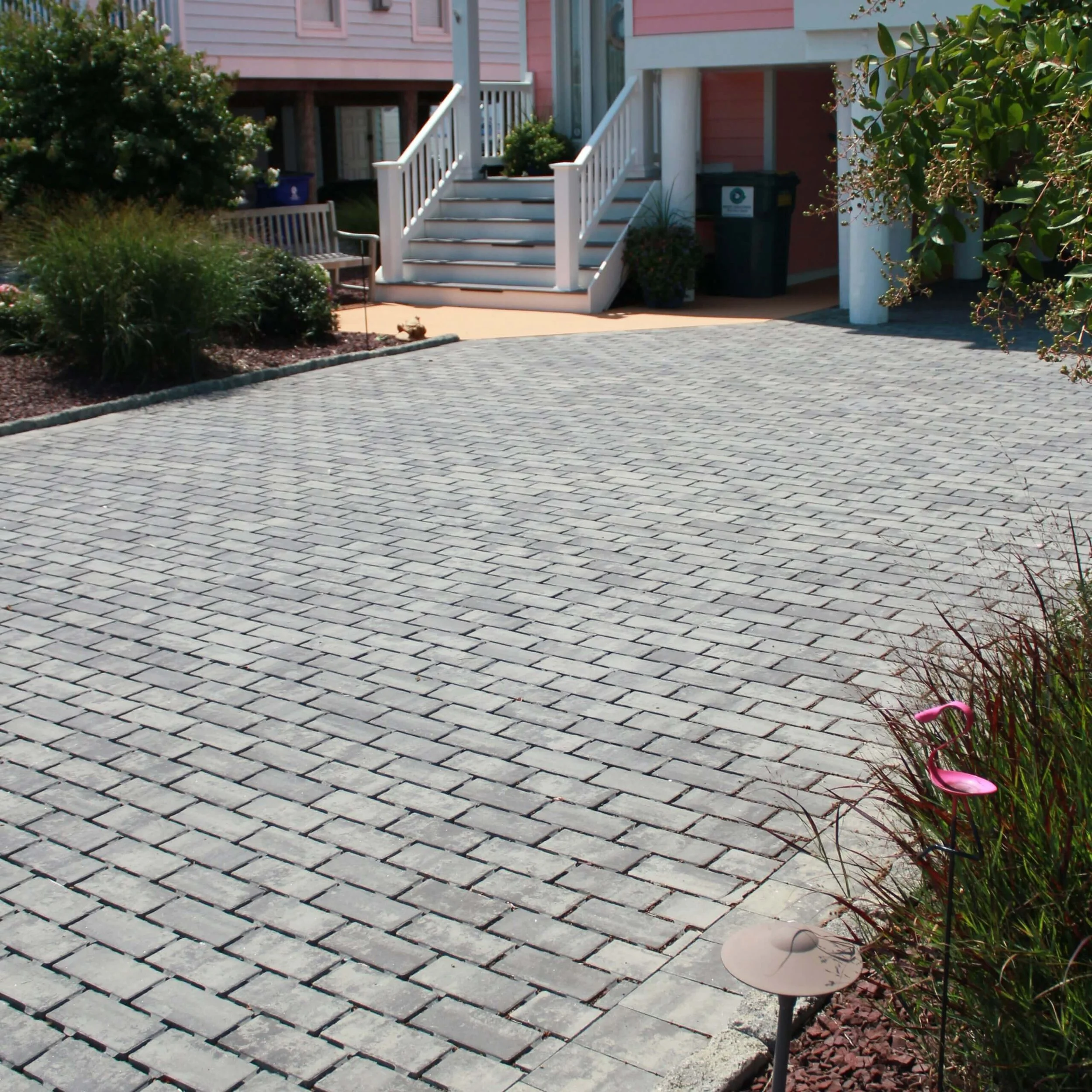 View of a paved driveway leading to a house with a pink exterior, white steps, and railing. There are plants and pink flamingo lawn ornaments on the right side.