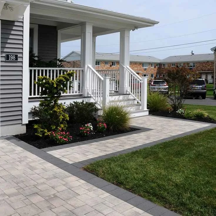 Front porch with white railing, stairs, and columns; landscaped garden beds with shrubs and flowering plants; paved walkway; neighboring houses and parked cars in background.