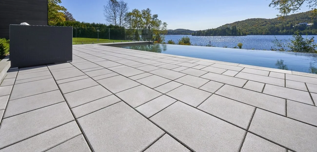 Photo of a pool deck, covered in a light gray Beacon Hill concrete paver, surrounding an infinity pool overlooking a woodland lake.
