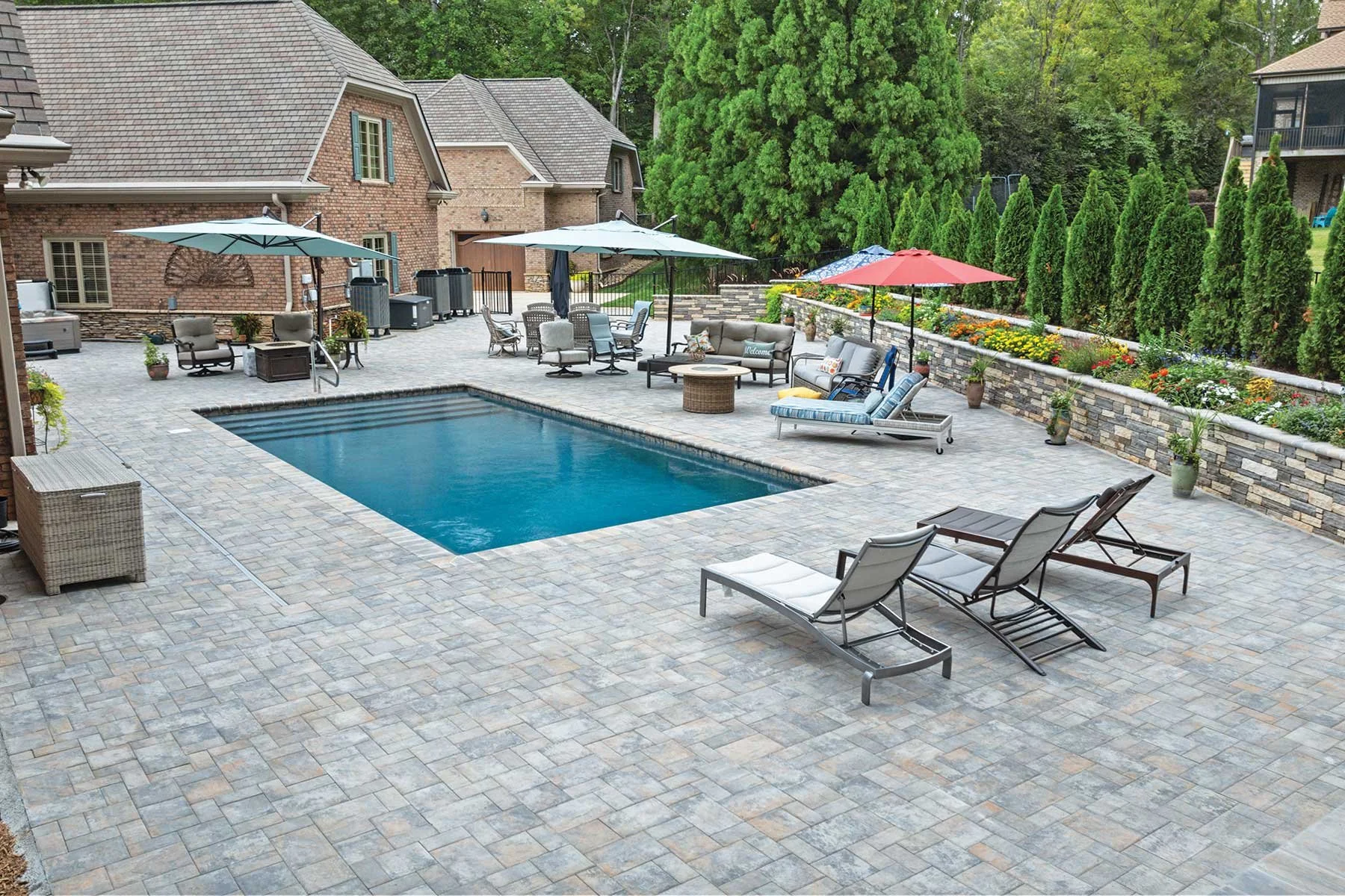 Backyard with a rectangular swimming pool, Nicolock concrete paver patio, outdoor furniture, umbrellas, a flower bed, trees, and a brick house.