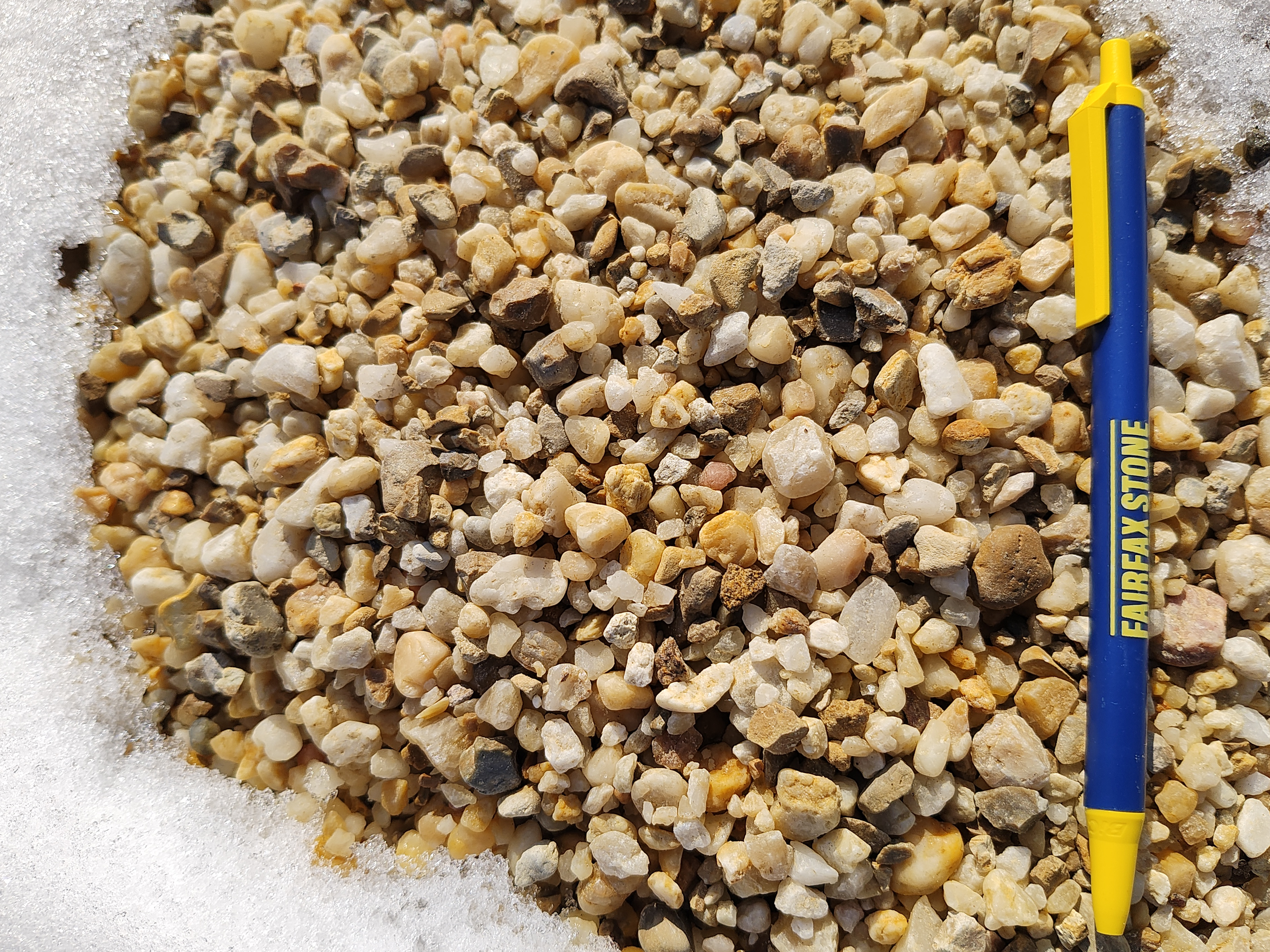 A pile of small, multicolored gravel pea gravel in snow with a blue and yellow Fairfax Stone marker pen lying across the gravel.