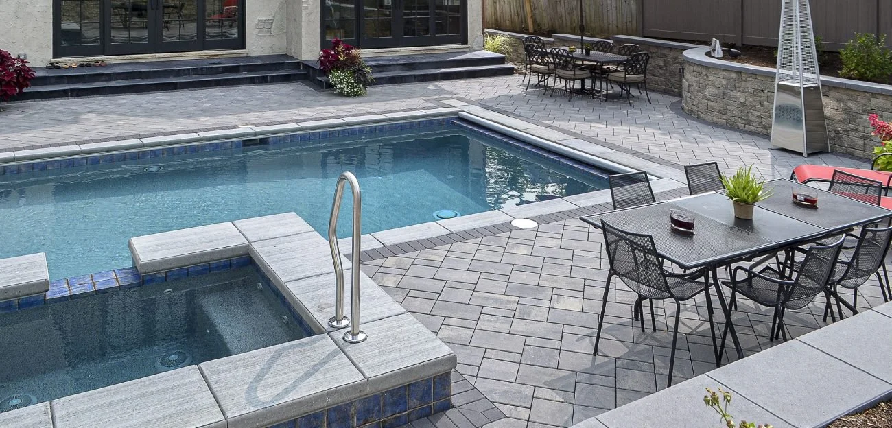 Two-tone gray Treo paver pool deck, bordered by a solid charcoal soldier course. A hot tub in the foreground empties into the main pool.