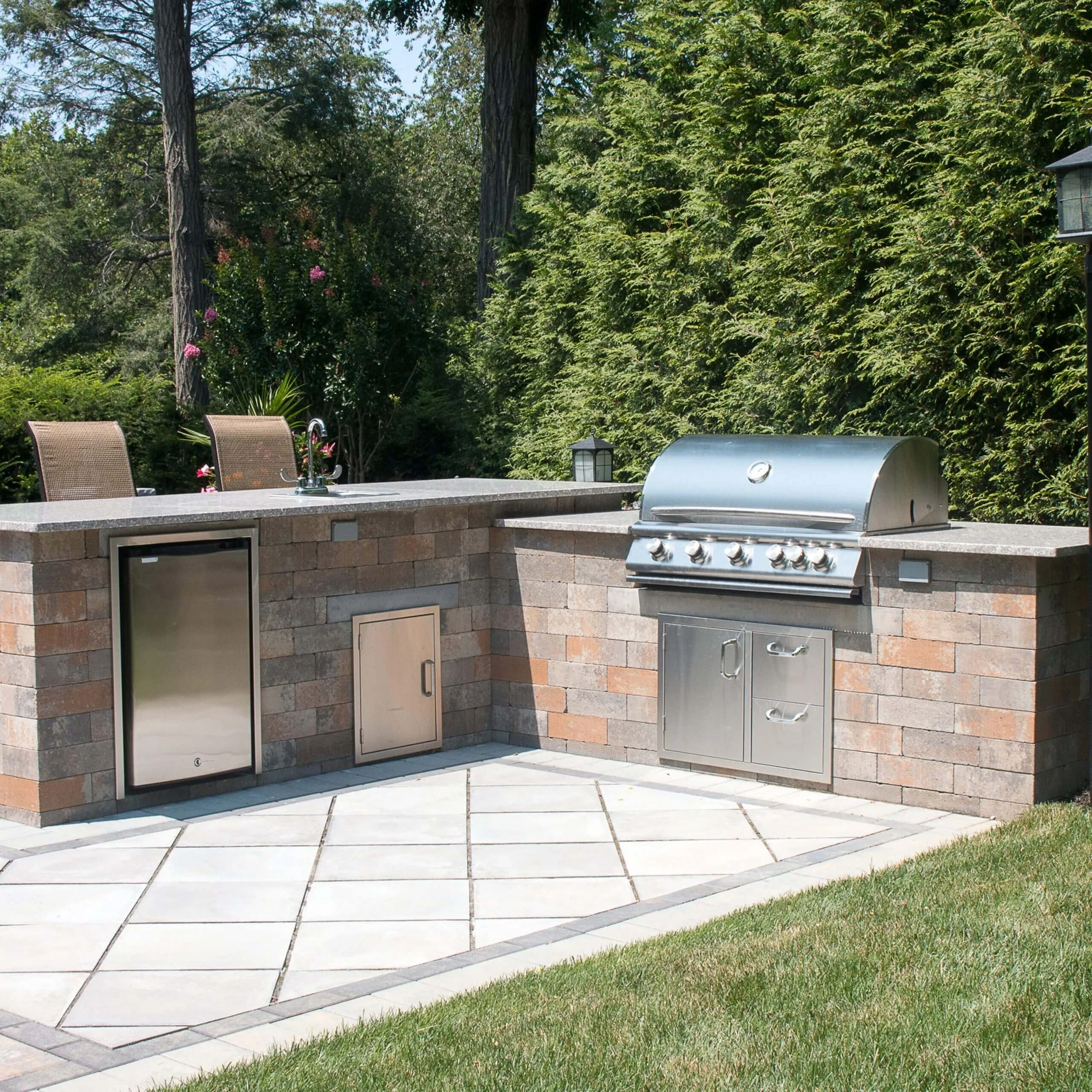 Outdoor kitchen with stainless steel grill built into cabinets built from Nicolock wall bloc.  Two chairs with greenery in the background.