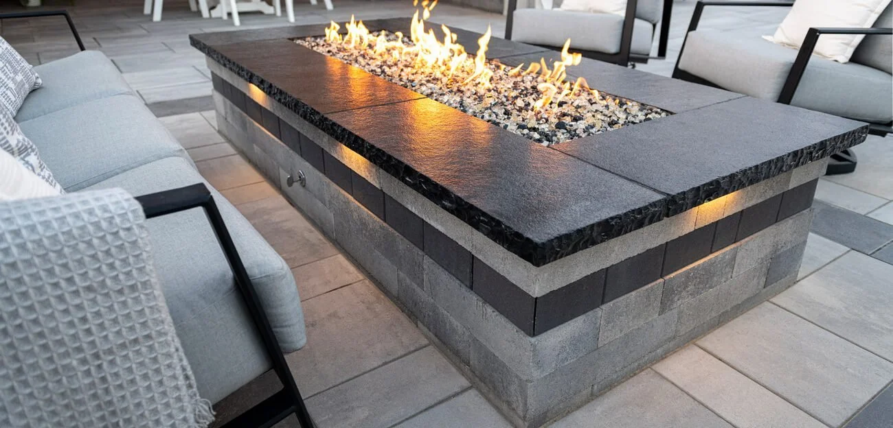 Custom firepit, built from Lineo wall using Granite wall with Graphite banding, a black limestone cap, and a linear gas insert.  Fire pit is the centerpiece of an outdoor seating area on a paver patio.