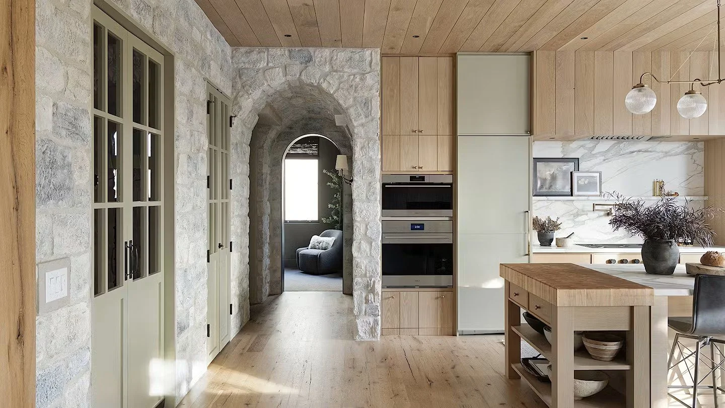 A modern kitchen with light wood cabinetry, a marble backsplash, and a kitchen island with open shelves. There is a gray refrigerator and an oven built into the cabinets. To the left, there is a stone wall with paneled doors and a large arched doorway leading to a sitting area with a gray armchair and window.