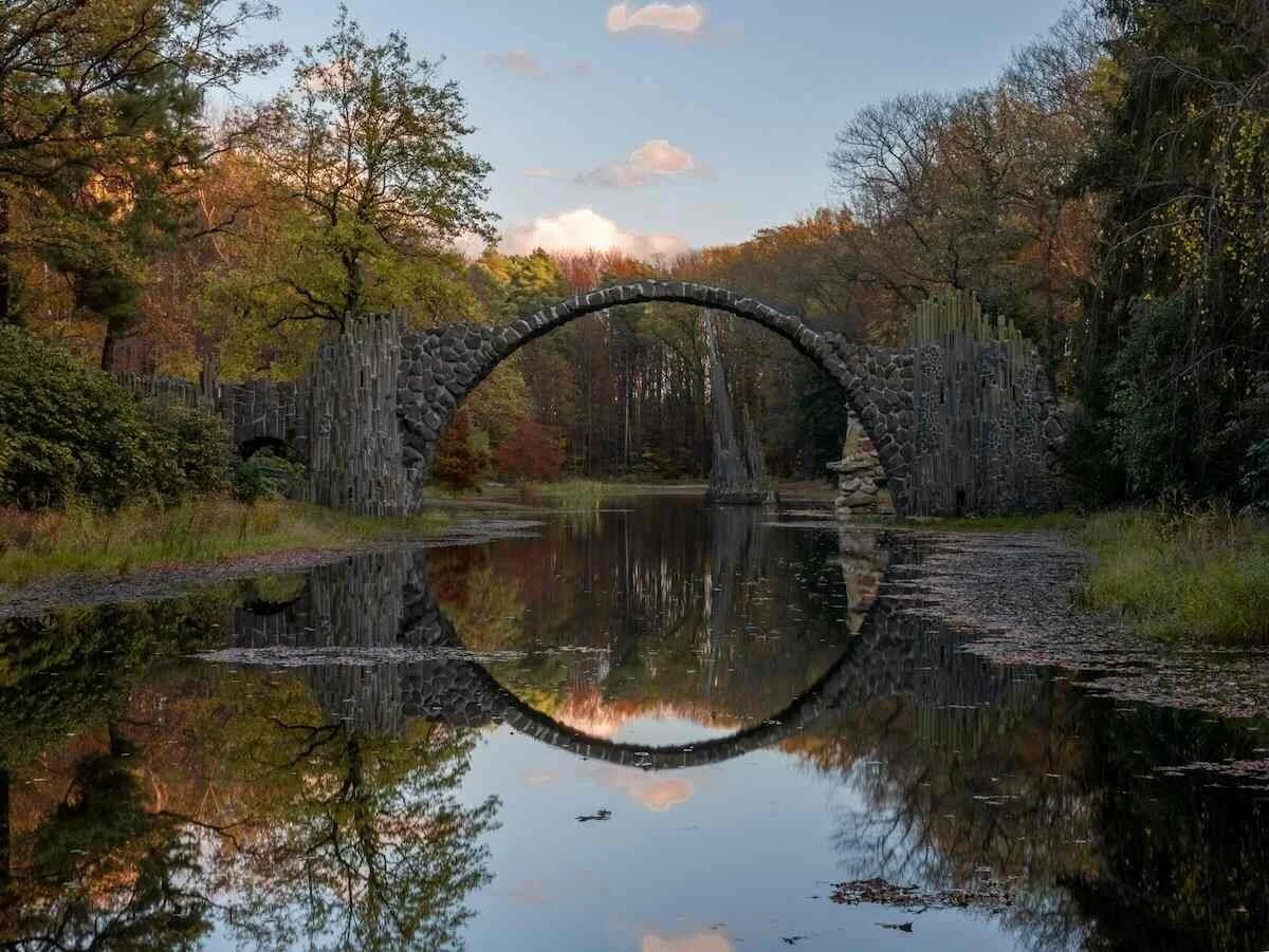 A stone arch bridge over a calm river, with trees showing autumn foliage and their reflection in the water.