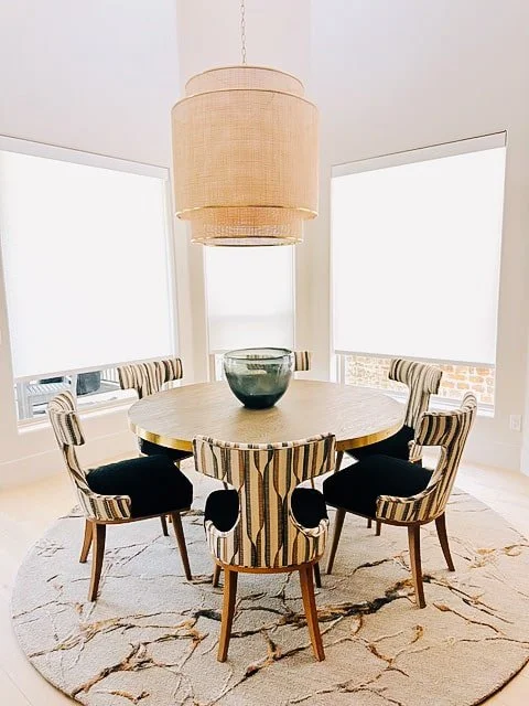 Round wooden dining table with a large bowl on top, surrounded by six striped upholstered chairs, in a bright room with large windows and a hanging pendant light