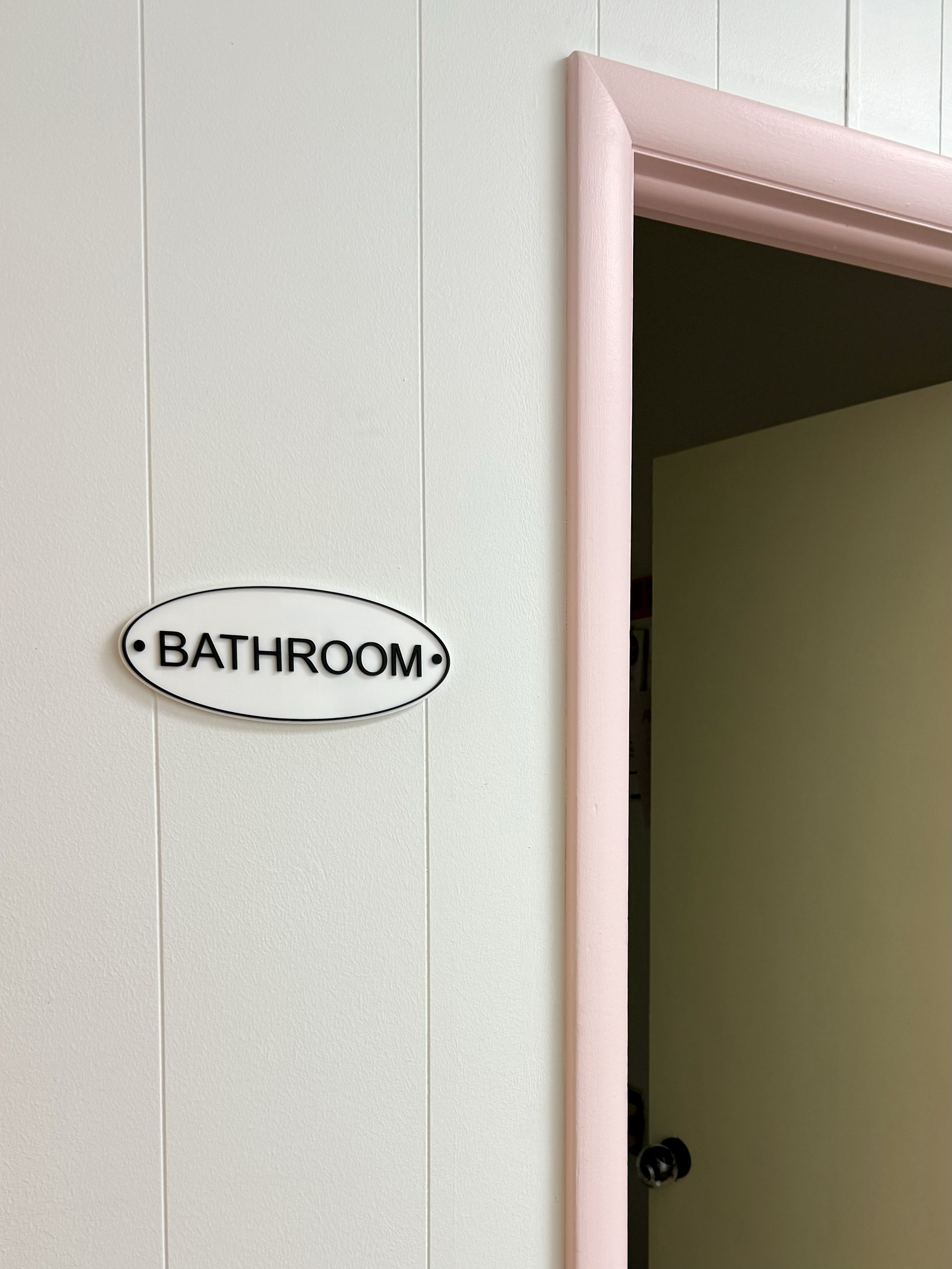 Bathroom sign next to an open door with a pink frame on a white wall.