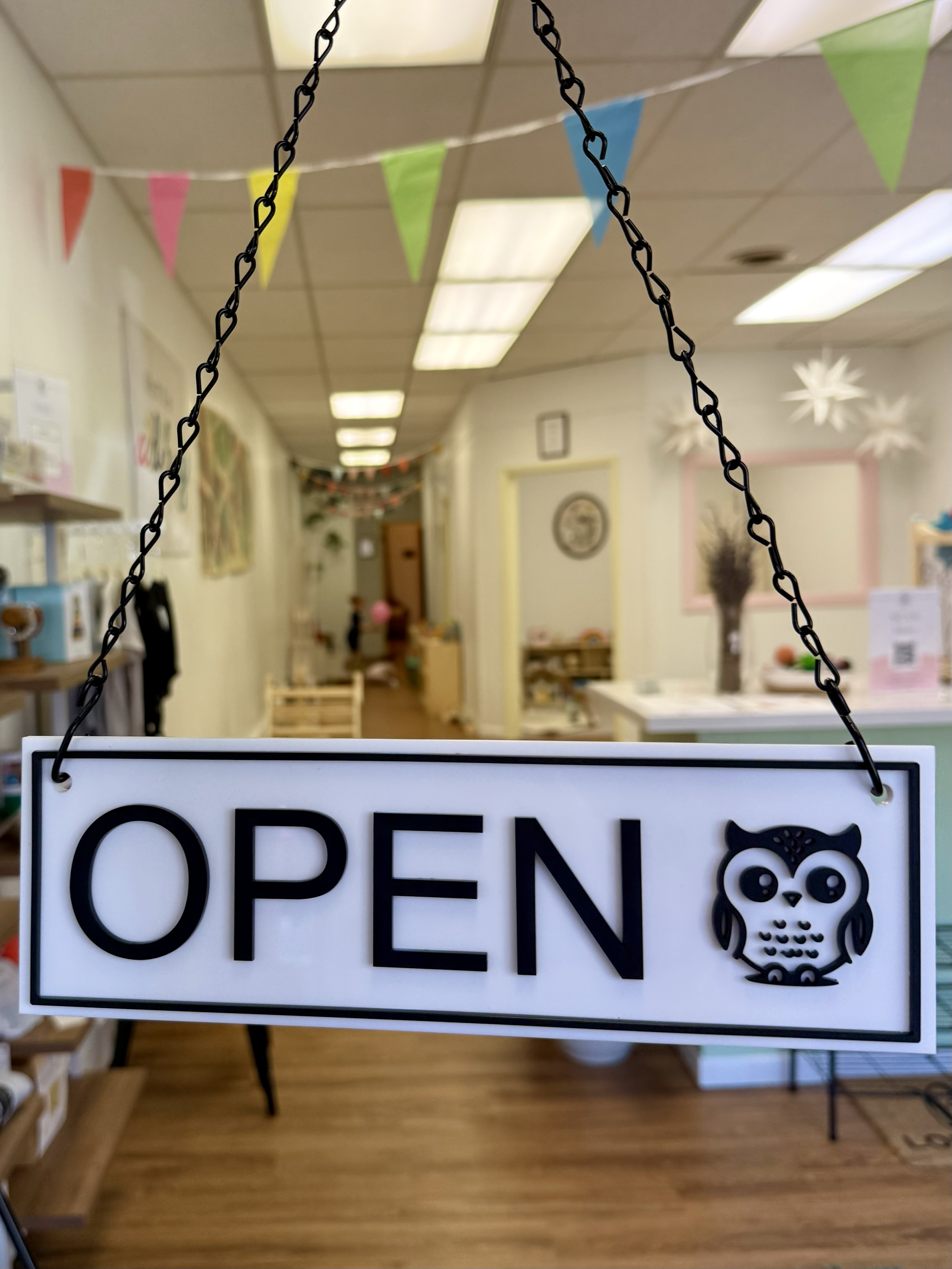 Open sign hanging on chain with owl illustration in a store interior decorated with colorful bunting and paper snowflakes.