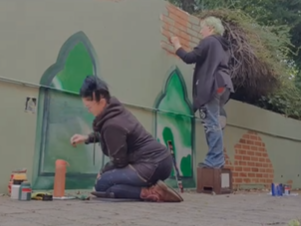 Two people painting a mural of green trees on a wall outdoors.