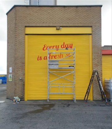 A yellow garage door with red painted words partly covered by a scaffold, reading 'Every day is a fresh start' with paint supplies nearby.