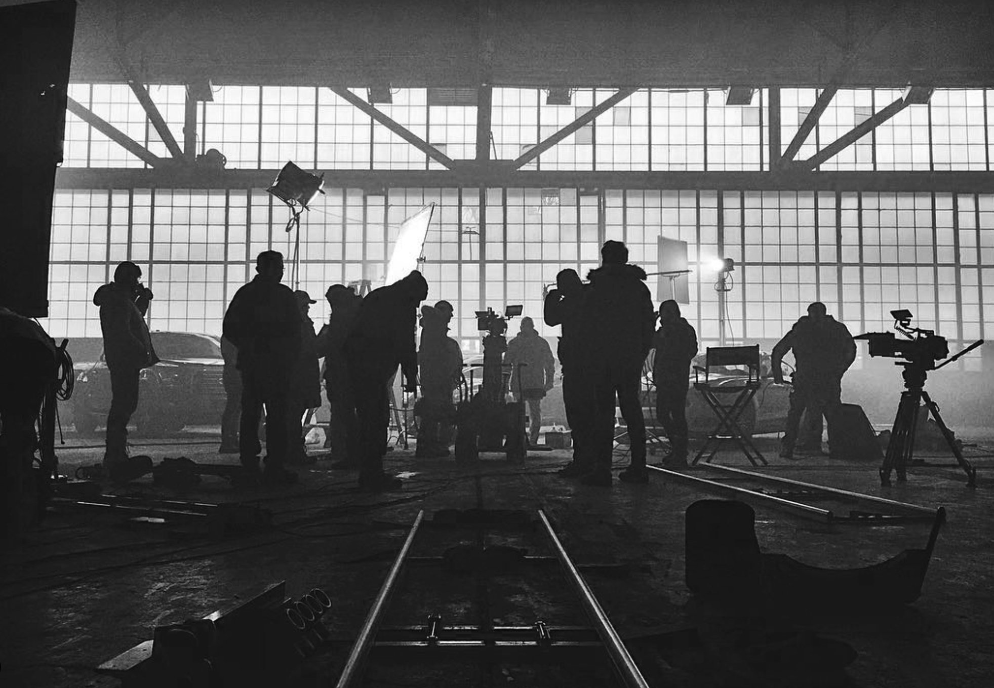 Black and white photo of a film or video production set inside a large industrial building with high windows, crew members, cameras, and lighting equipment.